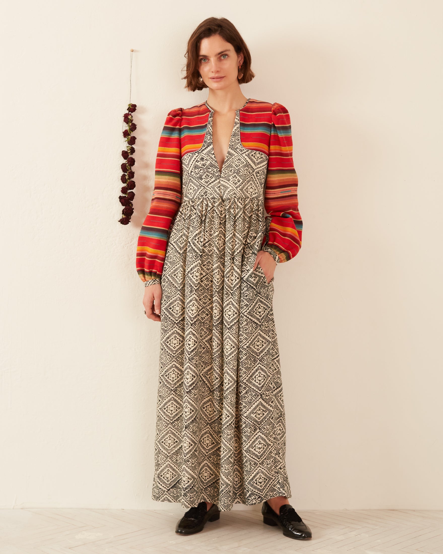 A woman stands against a white wall wearing the Winifred Orange Bloom Dress, featuring a gathered skirt and vibrant striped sleeves. Her hand rests in her pocket, and she's paired the outfit with black shoes. Beside her, a decorative hanging adorned with dark beads complements the dress's traditional craftsmanship.