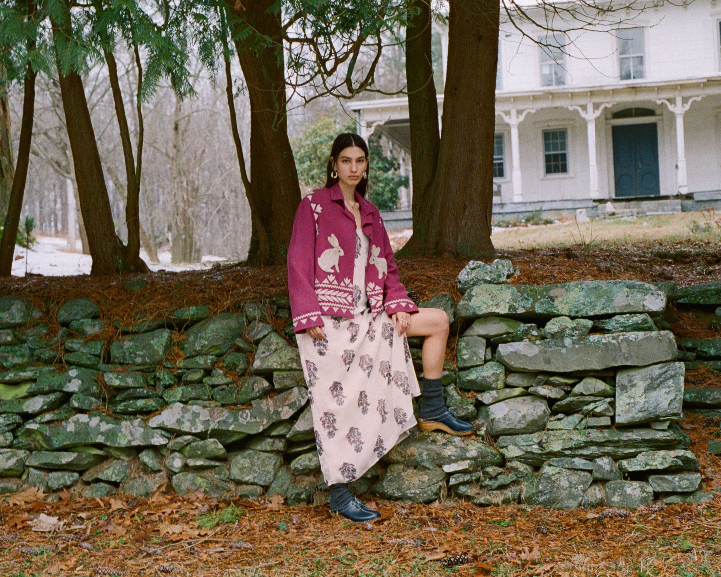 A person wearing a purple jacket and patterned skirt leans against a stone wall, surrounded by trees. A large, white house is in the background on an overcast day.