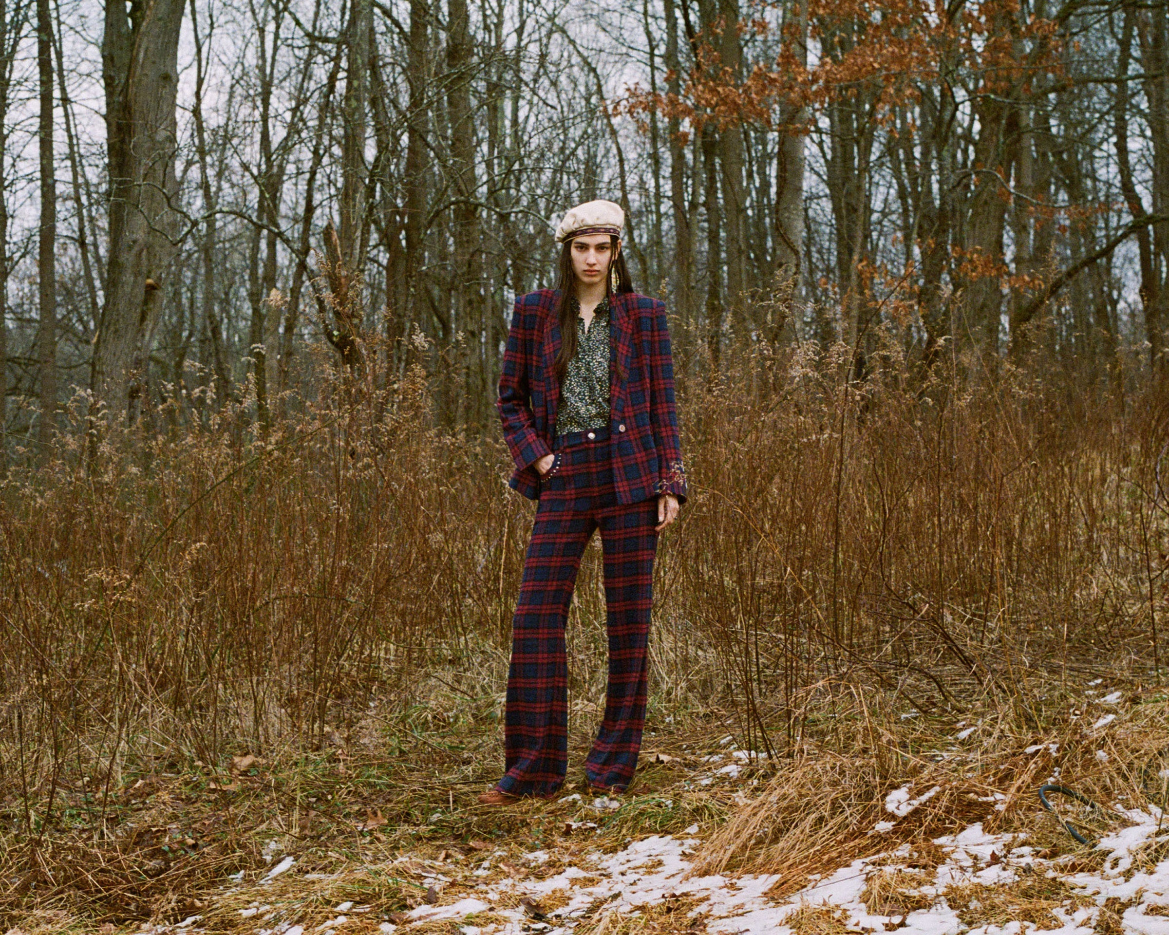 A person wearing a plaid suit and a beret stands in a grassy, wooded area. The ground is scattered with patches of snow, while the trees are bare, suggesting a late autumn or early winter setting.