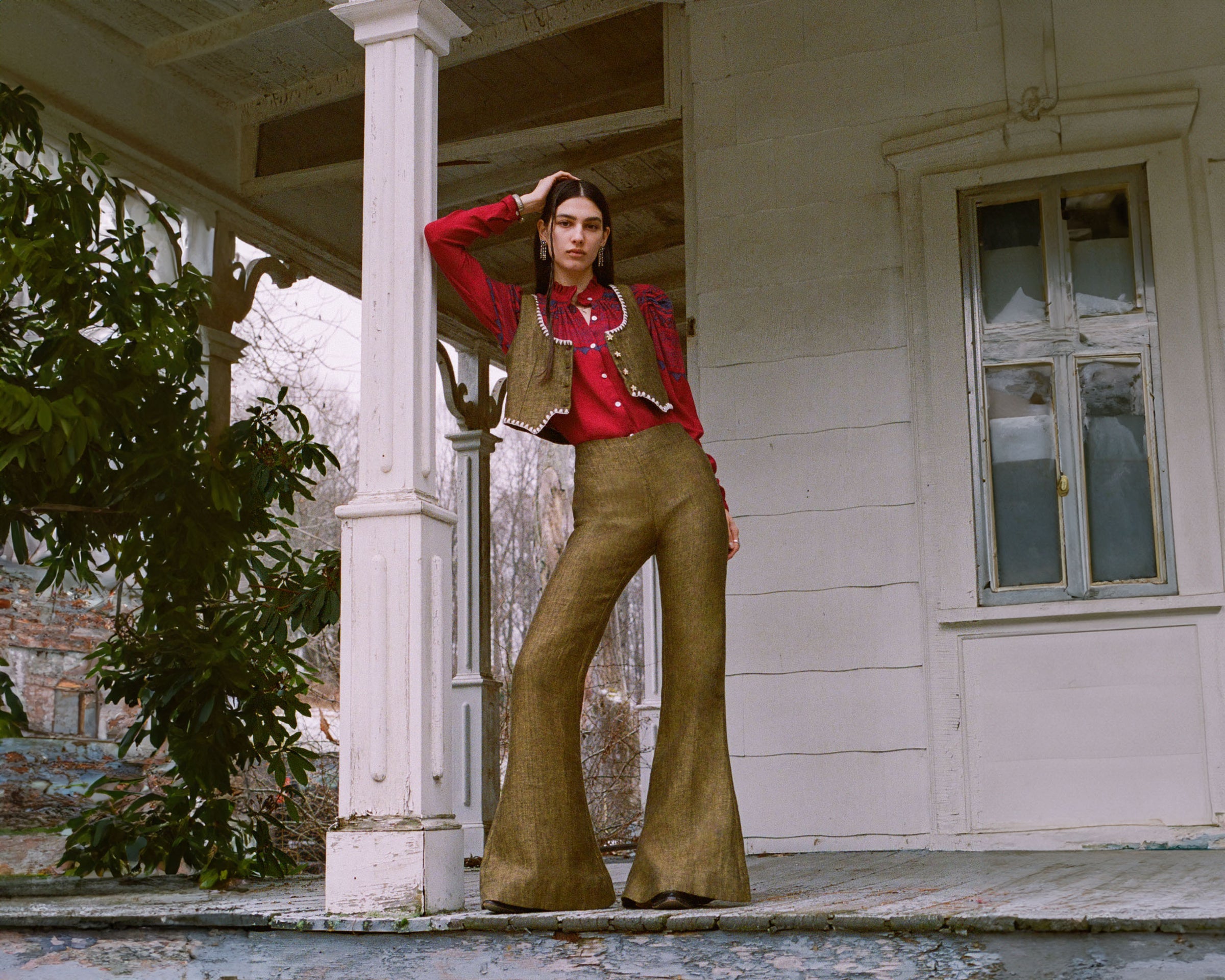 A person poses on a weathered porch wearing a red shirt, vest, and wide-legged trousers. They lean against a column with one hand on their head. The background features trees and an old, white building with a window.