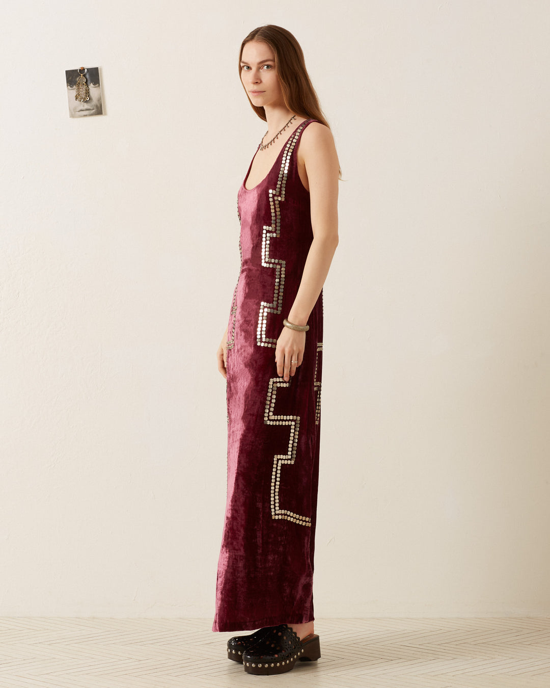 A woman stands against a plain wall wearing the Adele Plum Velvet Dress, a sleeveless piece adorned with geometric patterns. She looks directly at the camera, her long brown hair cascading down as she stands in black shoes. A small framed photo is visible on the wall beside her.