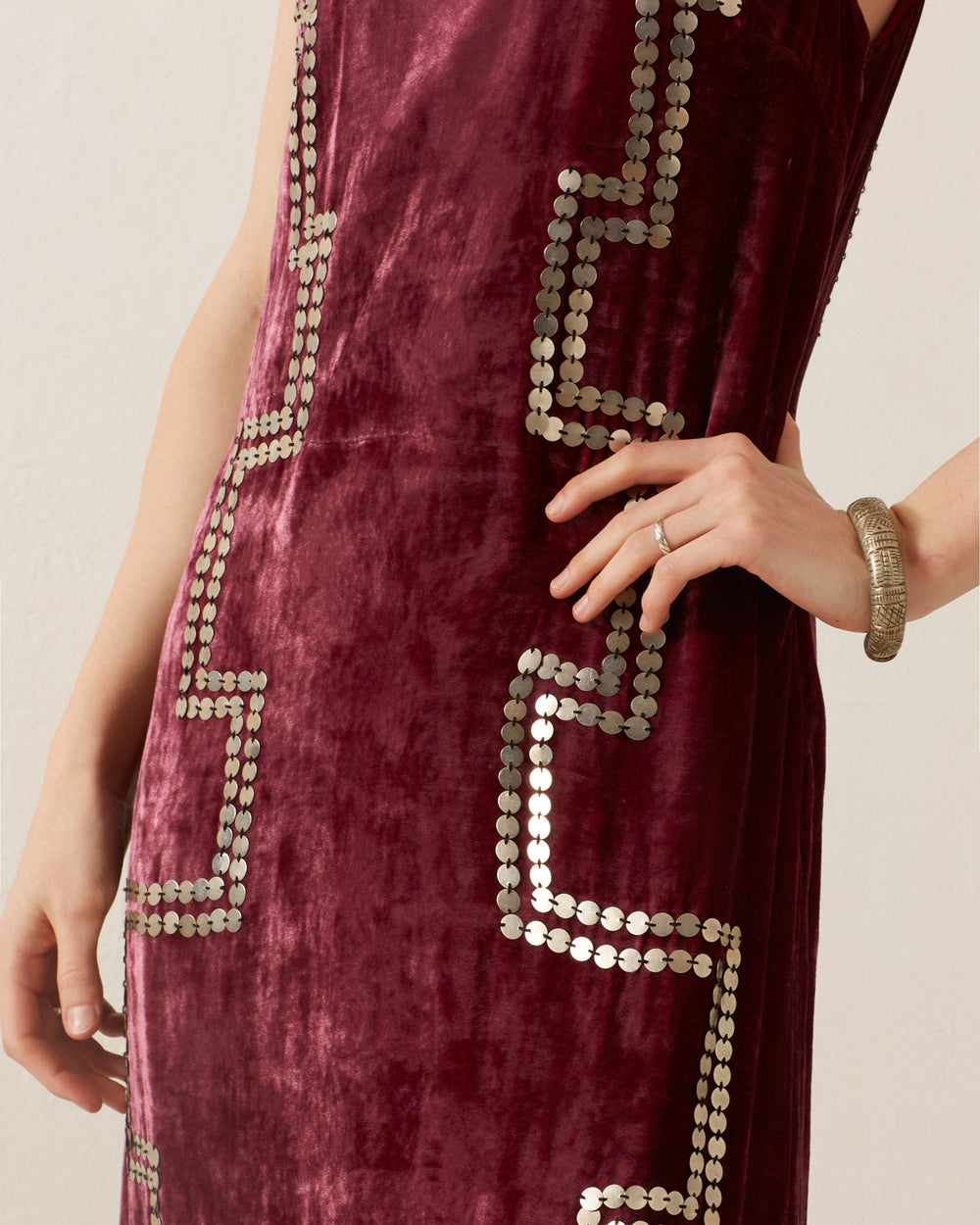 Close-up of a person wearing the Adele Plum Velvet Dress, featuring a sleeveless design in deep maroon with an intricate geometric pattern and zigzag silver embellishments. One hand is on their hip while the other rests by their side, showcasing a silver bracelet.