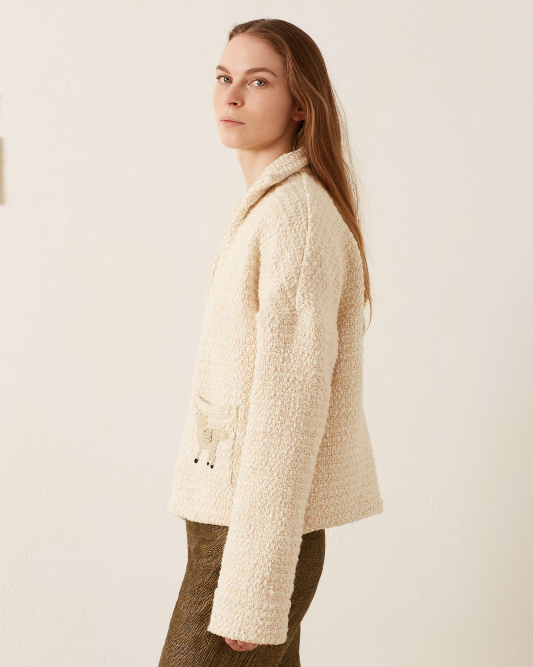 A person wearing the Allegra Pearl Boucle Top, a cream-colored sweater featuring hand embroidery, stands sideways against a plain white background, gazing forward.