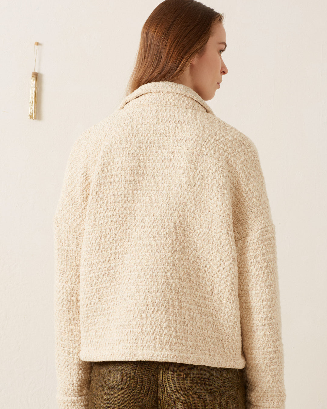 A woman stands facing away, wearing the Allegra Pearl Bouclé Top in cream paired with brown pants, set against a plain light background.