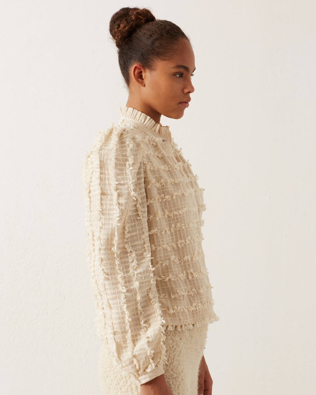 A person with a bun hairstyle stands sideways, wearing the Ana Snow Feather Shirt, a textured, cream-colored blouse featuring an elegant ruffled collar, paired with a matching skirt. The background is a plain, light-colored wall.