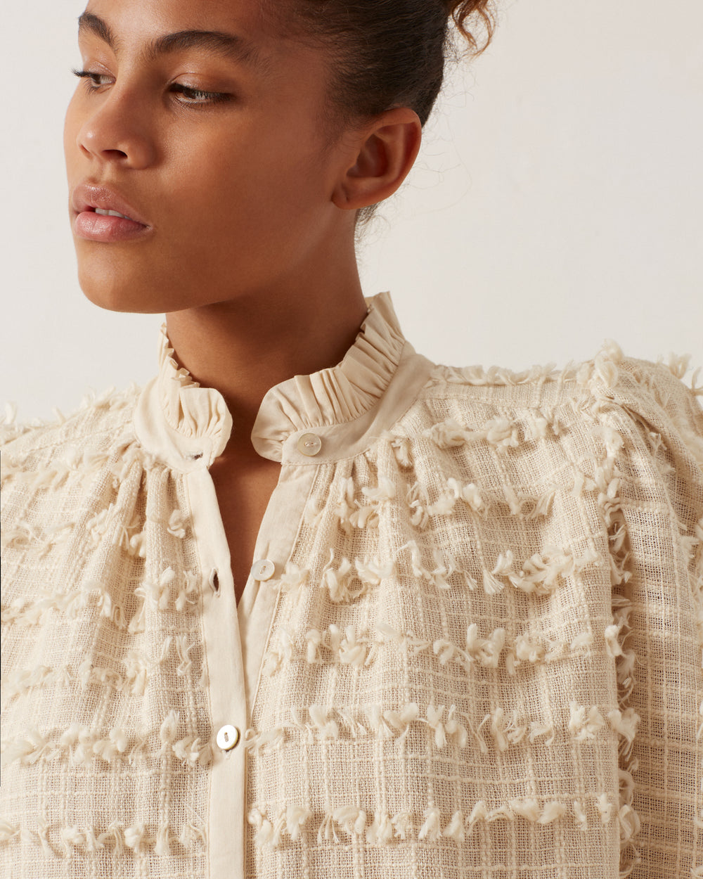 A person wearing the Ana Snow Feather Shirt, which features an ivory color with a high ruffled collar and buttoned front, is looking to the side against a light background.