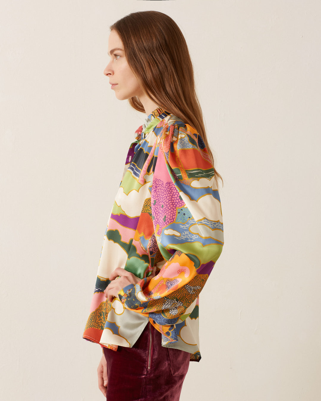 A woman with long brown hair is standing sideways, elegantly wearing the Annabel Aurora Silk Shirt, featuring a colorful Art Nouveau landscape print, paired with burgundy pants against a plain background.