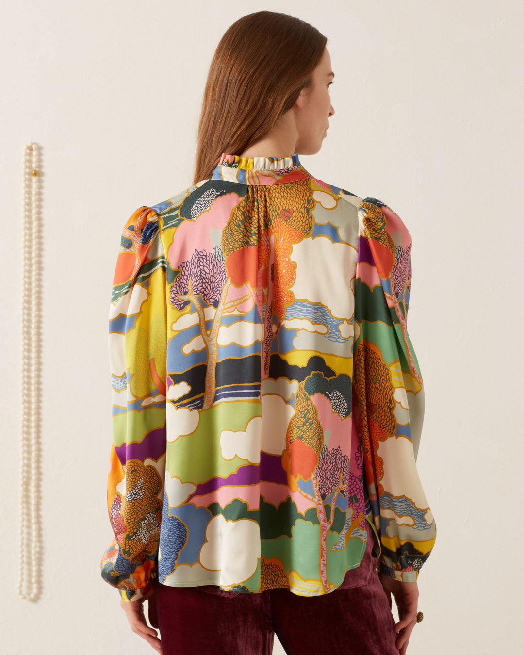 A woman with long brown hair wears the vibrant Annabel Aurora Silk Shirt, featuring a Liberty print and a high collar, as seen from behind. She stands against a plain, light-colored wall, with a string of pearls elegantly draping down.