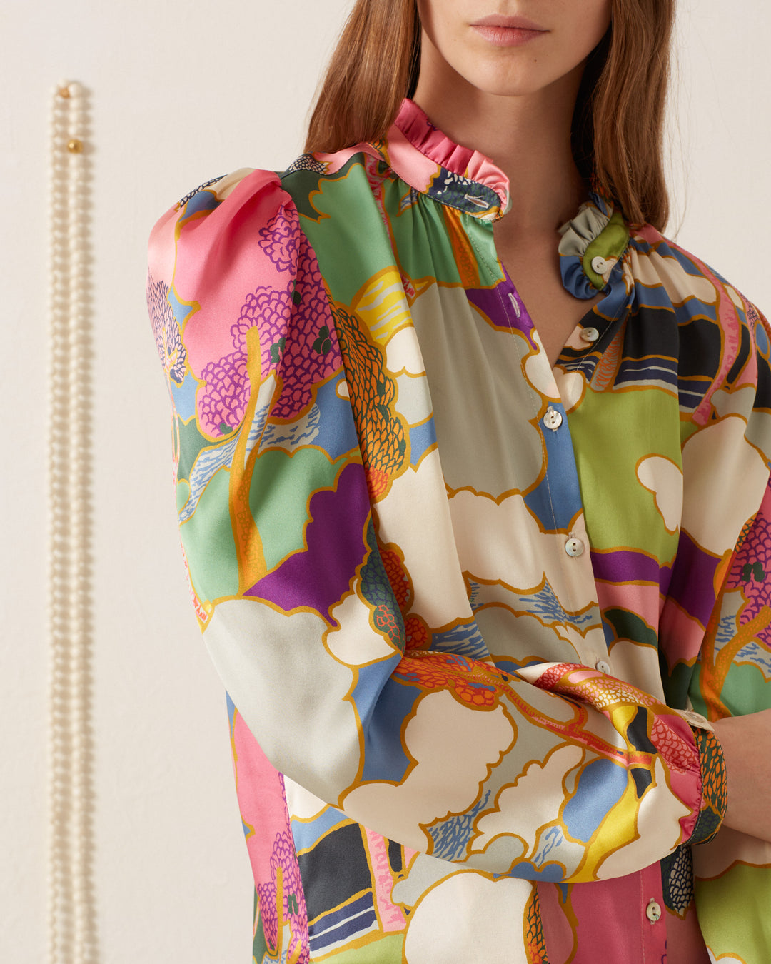 Wearing the vibrant Annabel Aurora Silk Shirt with a ruffled collar, an individual stands against a white wall, where the blurred elegance of an Italian silk charmeuse necklace is visible in the background.