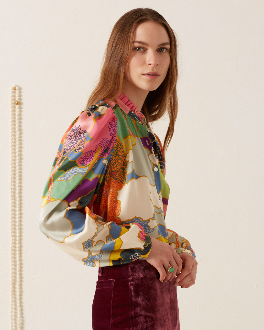 A woman in a vibrant Annabel Aurora Silk Shirt and maroon skirt stands gracefully against a light background.