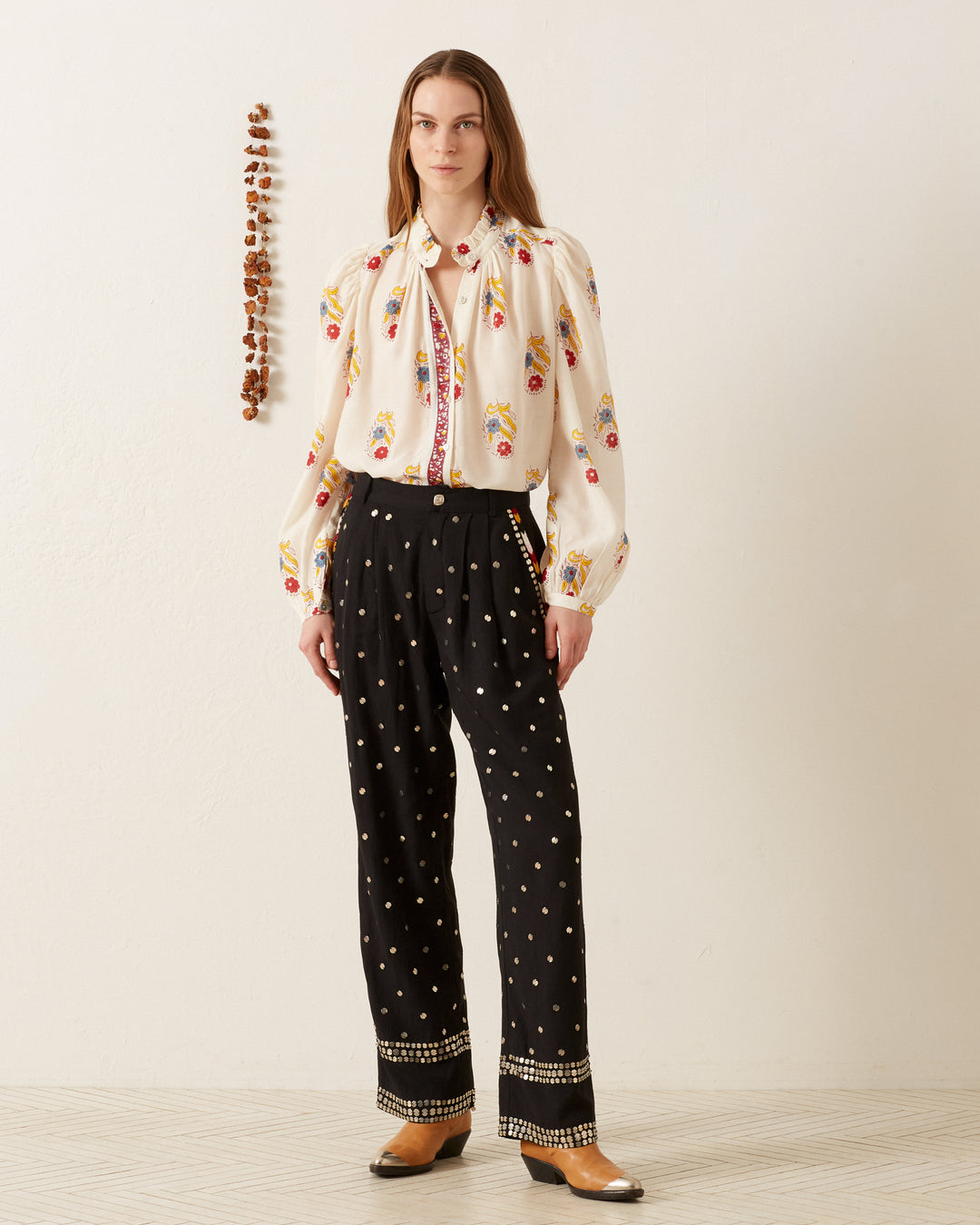 A person stands against a beige wall wearing the Annabel Fireflower Shirt, a white blouse with colorful embroidery, paired with black Tencel twill pants dotted with white. They have long hair and are wearing brown boots. A string of beads hangs on the wall.