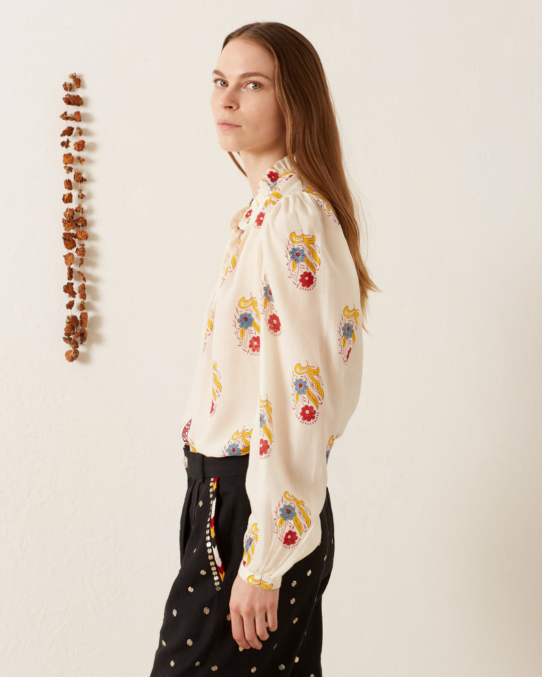 A person with long brown hair is wearing the Annabel Fireflower Shirt, featuring colorful floral motifs, paired with black polka dot pants. They stand in profile against a plain wall, accented by a hanging string of brown beads.