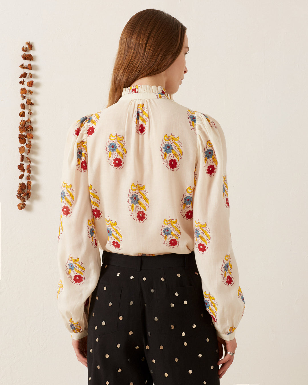 A woman with long brown hair faces away from the camera, wearing the Annabel Fireflower Shirt with its colorful floral hand-block print, paired with black pants adorned with white polka dots. The wall is adorned with a string of small brown objects.