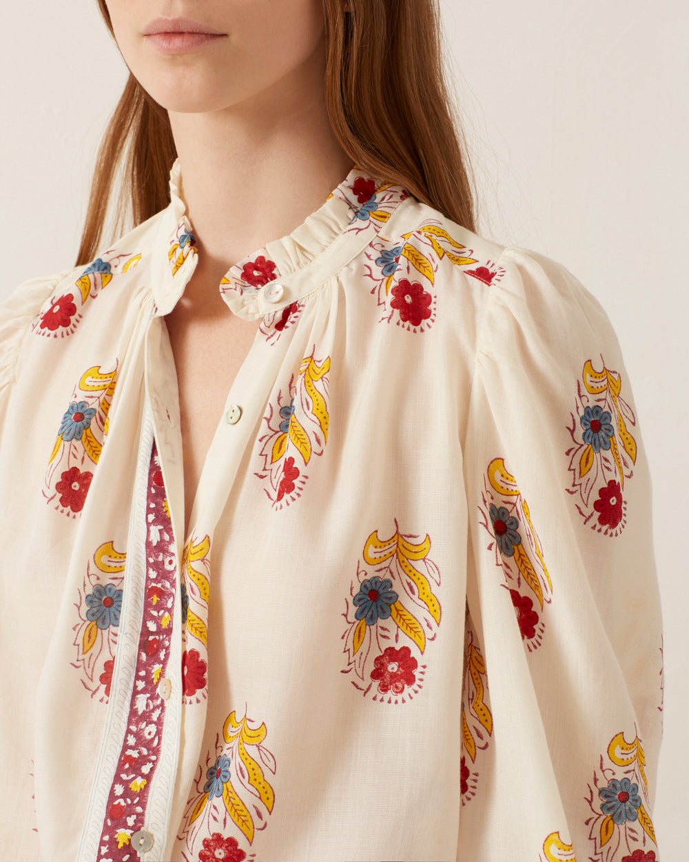 A woman wearing the Annabel Fireflower Shirt, featuring a cream-colored floral motif with vibrant red, yellow, and blue flowers. The blouse is accented with a ruffled collar and a partial button-up front. Her long, straight hair beautifully complements the delicate design.