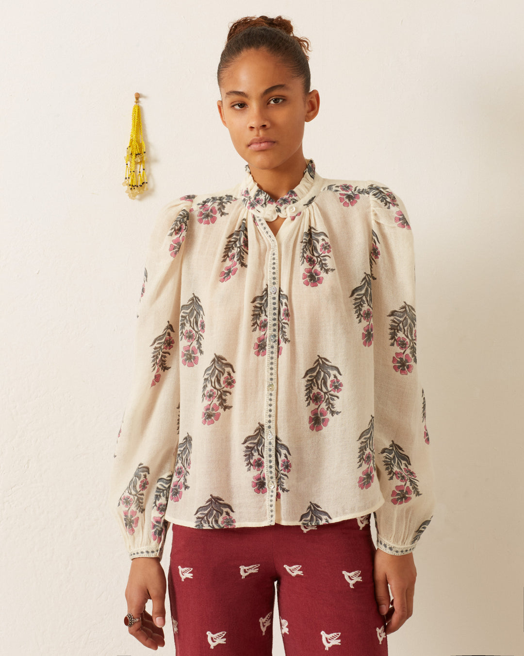 A person stands against an off-white wall, wearing the Annabel Mirabilis Wool Shirt with a floral motif paired with burgundy pants featuring bird designs. Their hair is tied up, and a decorative wall hanging is visible in the background, complemented by a hand-block printed scarf draped elegantly around their neck.