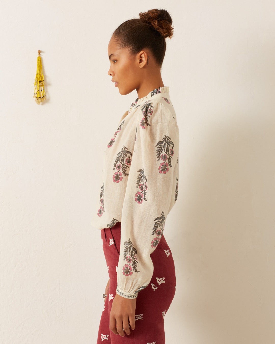 Someone with a bun hairstyle wears an Annabel Mirabilis Wool Shirt, which features a floral motif design, along with red pants adorned with butterfly prints. They stand sideways against a plain white wall, accented by a small decorative hanging in the background.