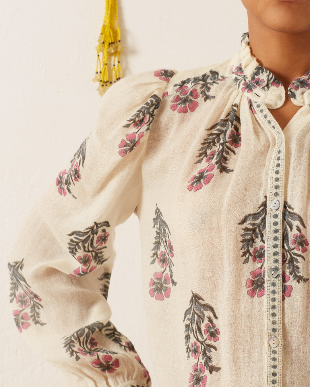 A person wearing the Annabel Mirabilis Wool Shirt, which is a cream-colored, hand-block printed blouse with a floral pattern in pink and gray. The blouse showcases a ruffled collar and button-down design. Yellow tassels hang on the wall in the background, with only part of the person's upper body visible.