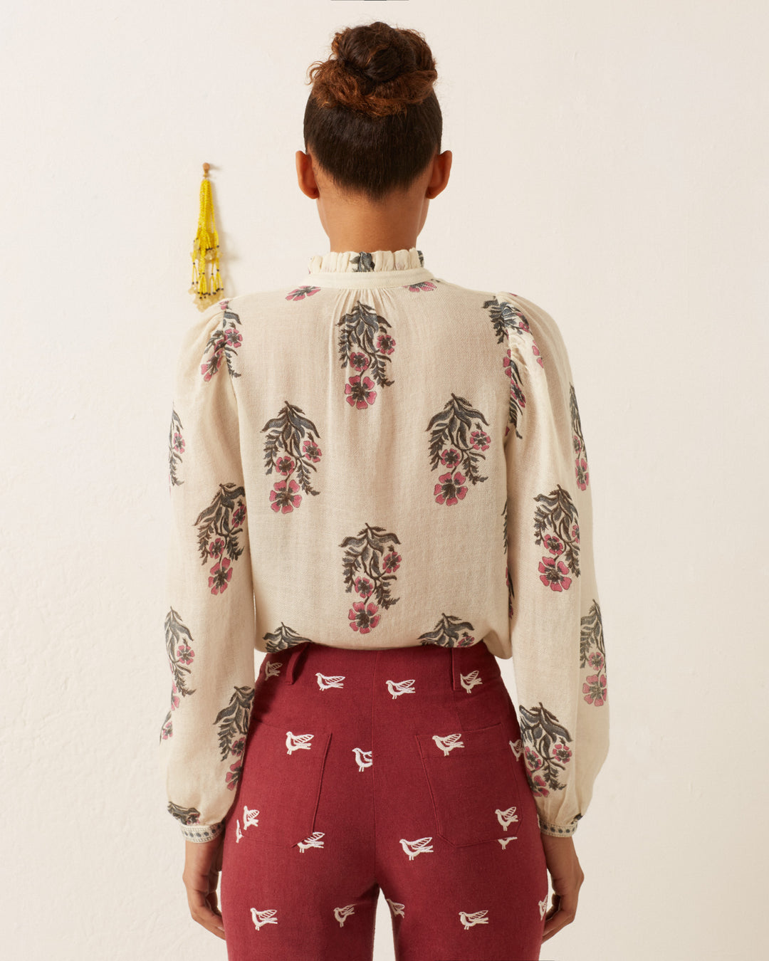 A person with short hair is standing with their back to the camera, wearing the Annabel Mirabilis Wool Shirt featuring a floral motif in cream tones, paired with maroon trousers adorned with bird patterns. A small decorative tassel hangs on the white wall, adding charm to the scene.