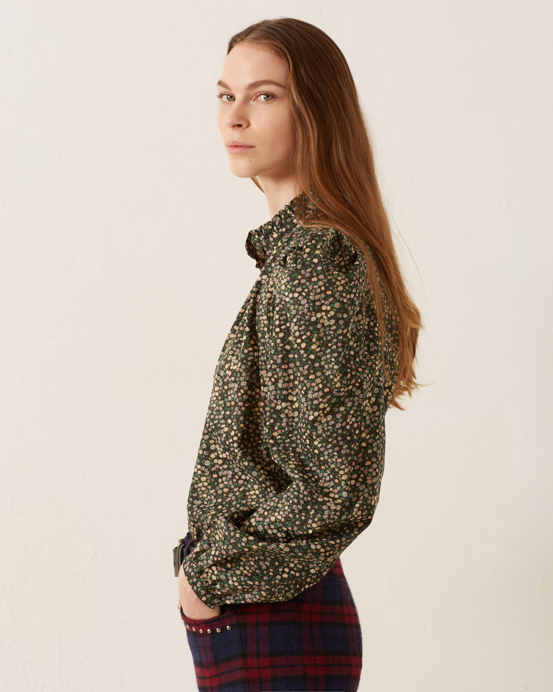 A person with long brown hair stands sideways, looking at the camera. They are wearing a dark Annabel Moonflower Shirt and a plaid skirt against a plain light background.