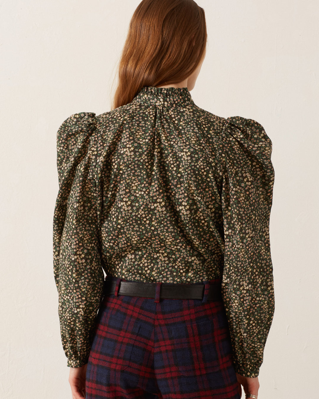 A person with long brown hair is wearing the Annabel Moonflower Shirt, a vintage-inspired blouse featuring a high neck and puffed sleeves, paired with dark plaid pants. The ditsy floral pattern elegantly drapes as they face away, showcasing the back details of the outfit against a plain light-colored background.