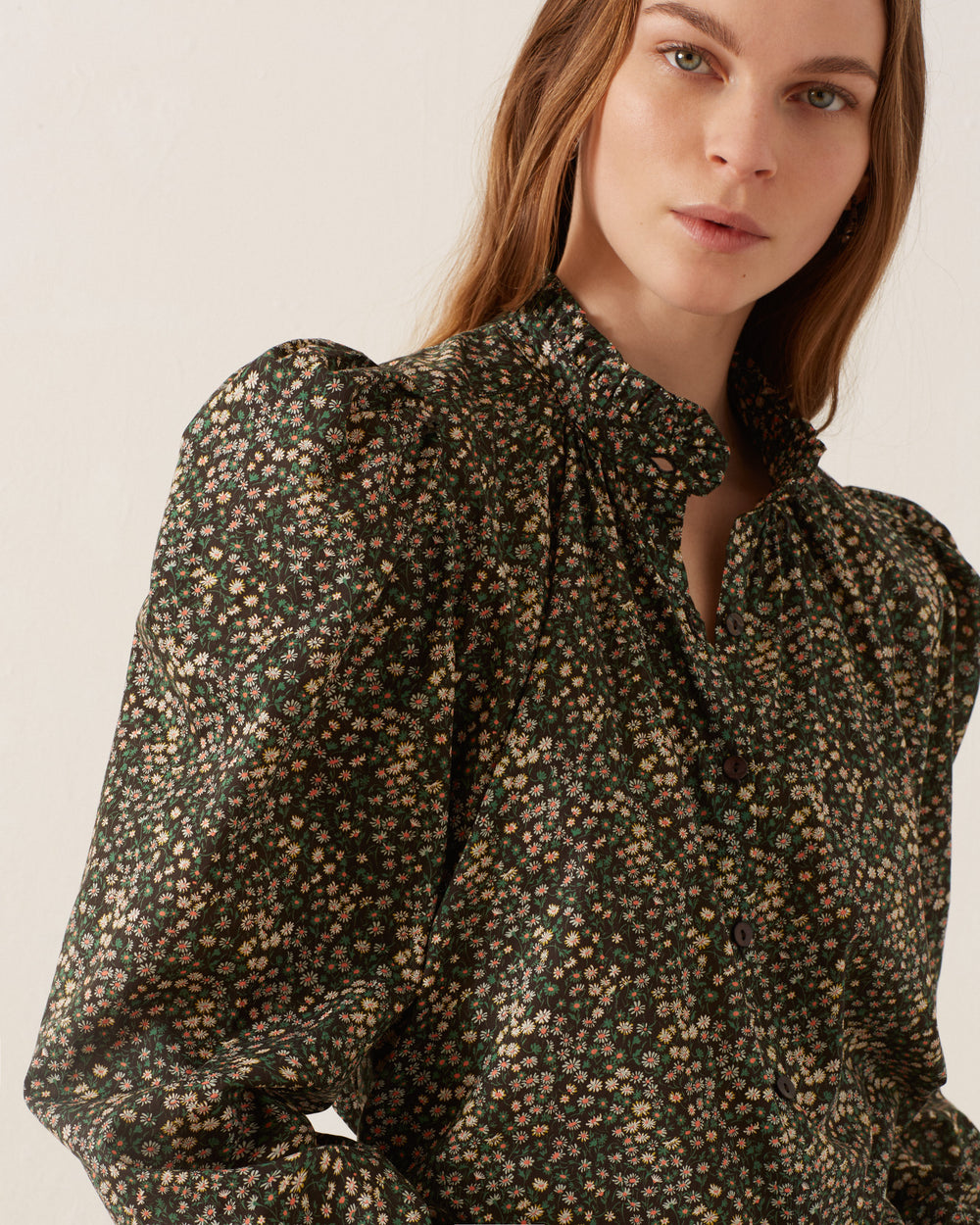 A woman with long hair wears the Annabel Moonflower Shirt, a vintage-inspired blouse with a dark ditsy floral pattern and puffed shoulders. She stands against a plain light-colored background, gazing directly at the camera.