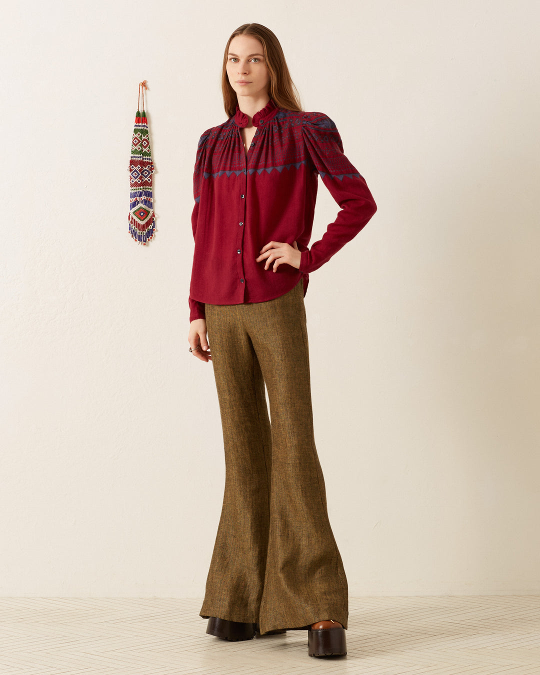 A person stands against a white background wearing an Artemis Plum Fairisle Shirt and brown flared pants. A colorful woven decoration hangs on the wall, adding vibrancy to the scene.