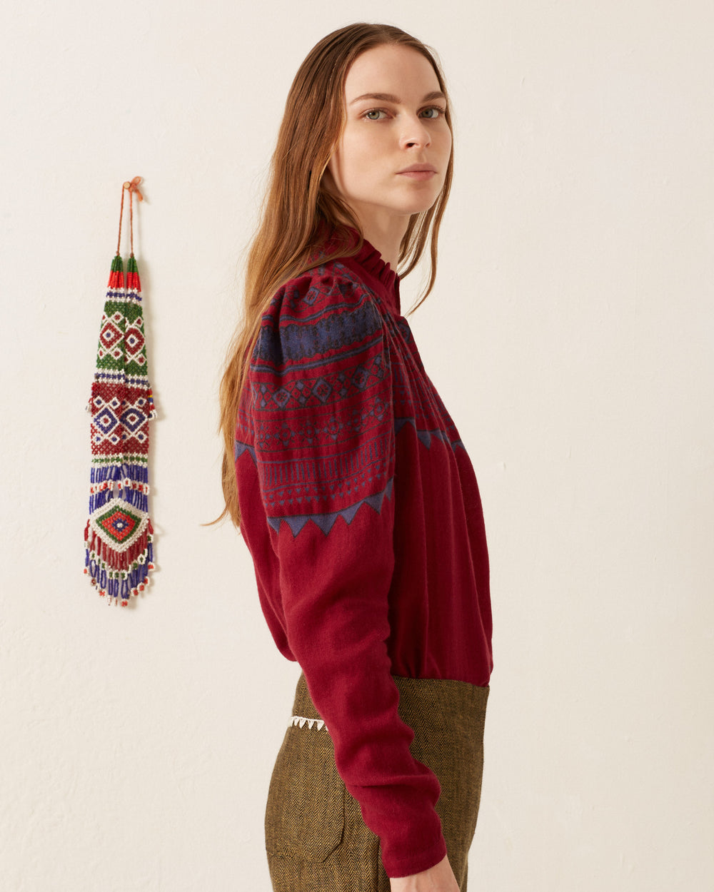 A person wearing an Artemis Plum Fairisle Shirt stands against a white wall, with a beaded wall decoration hanging nearby.