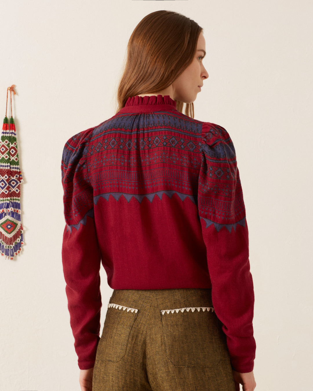 A woman with long hair is facing away, wearing the Artemis Plum Fairisle Shirt adorned with blue and white patterns, paired with brown pants. There is a beaded decoration on the wall.