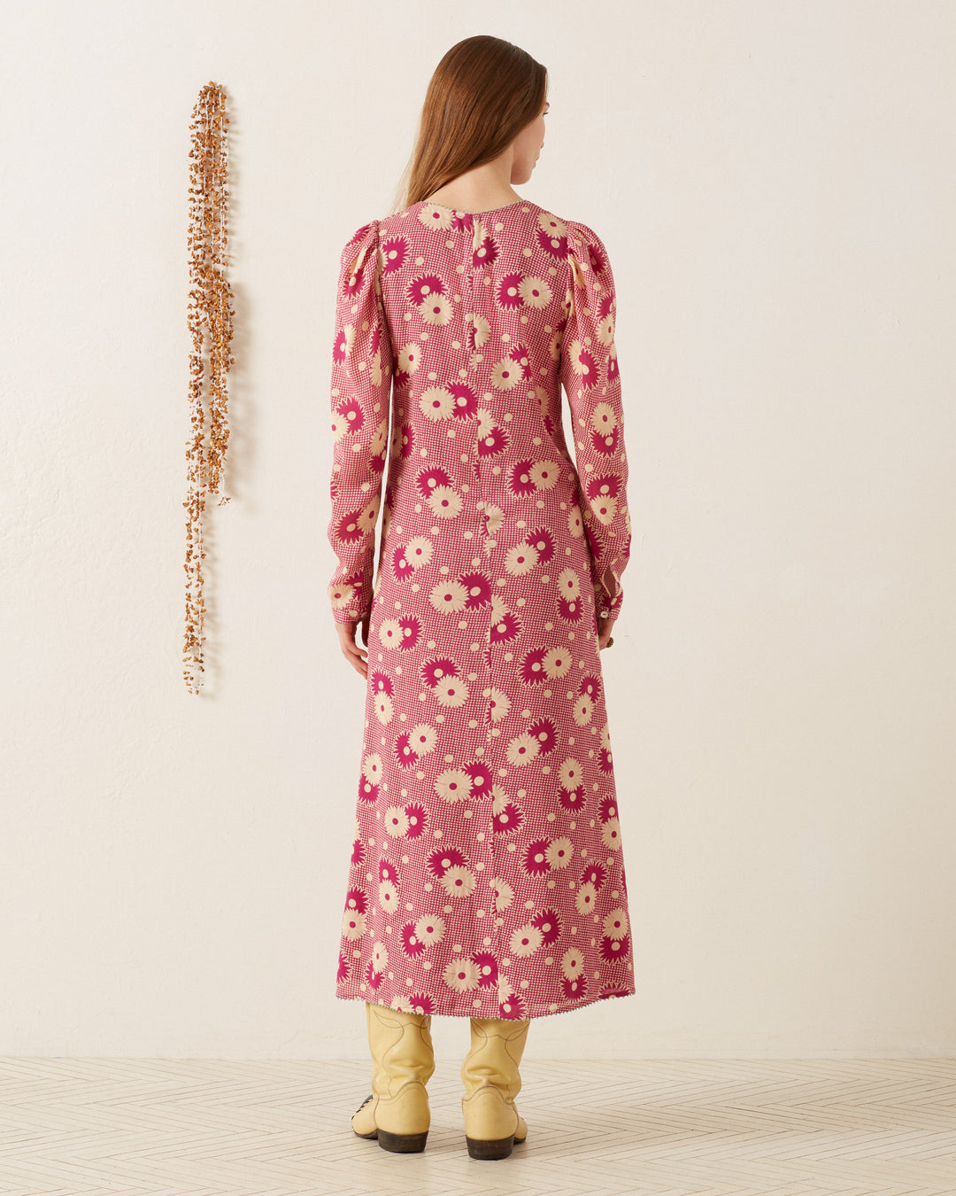 A person with long hair stands facing a light-colored wall, wearing the Astrid Berry Daisy Dress paired with beige boots. This vintage-inspired dress features puffed sleeves and a '70s-style floral print. To the left, a decorative beaded strand hangs on the wall.