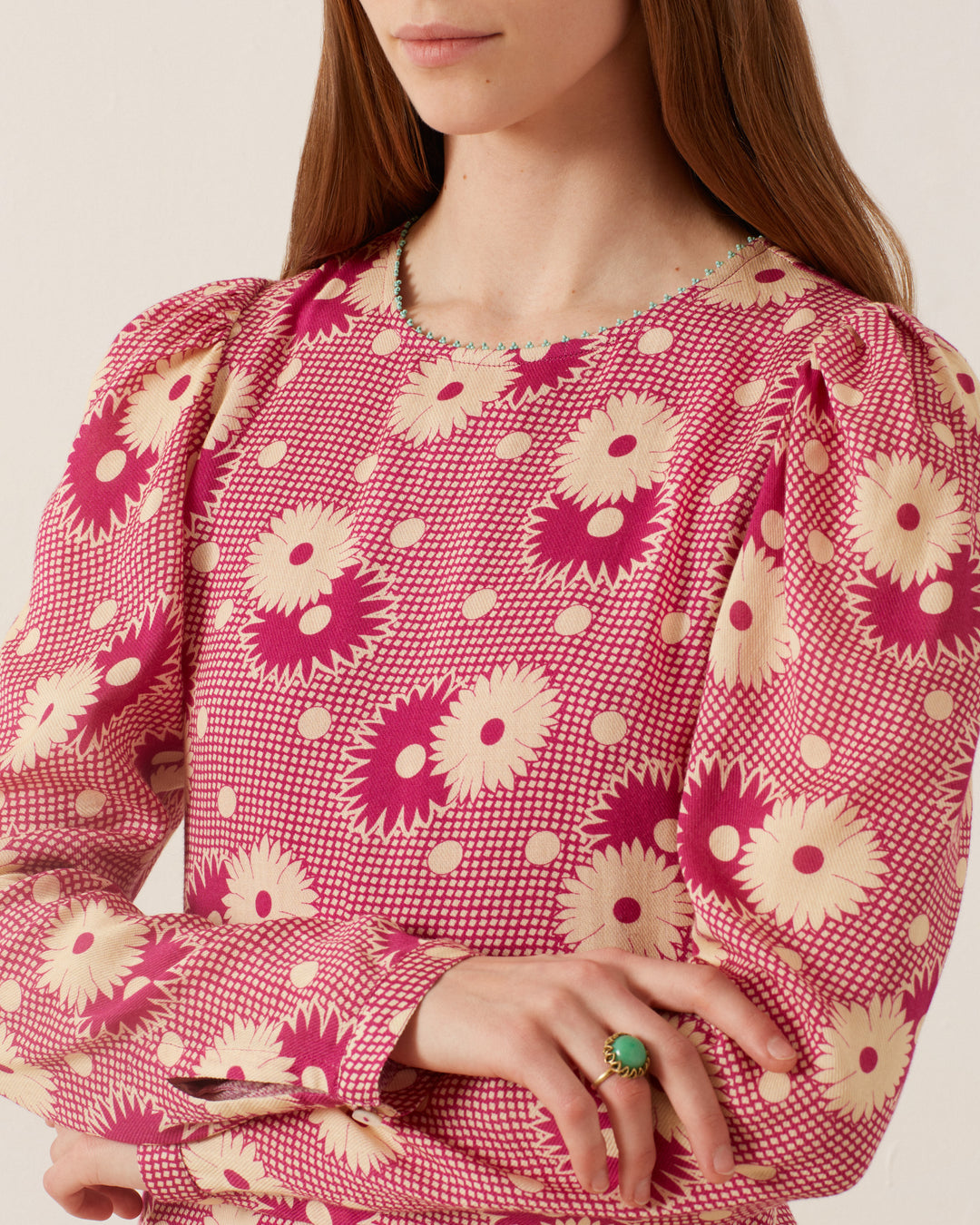 A person wearing the Astrid Berry Daisy Dress, a '70s-style garment featuring a floral print and puffed sleeves, complemented by a green ring on their finger. The dress showcases large flowers, polka dots, and a geometric design in pink and cream hues. The person's face is not visible.