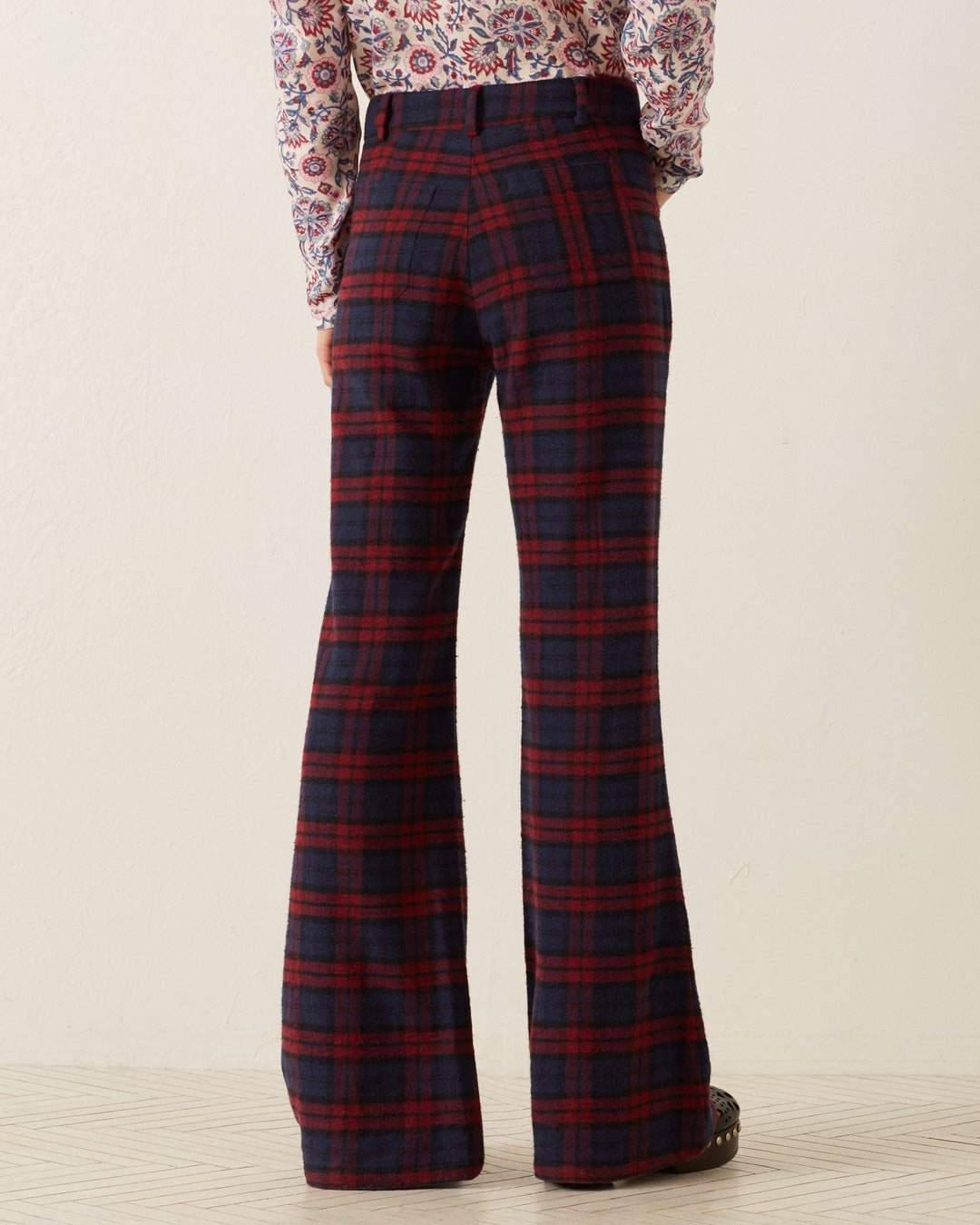 A person wearing the Bunny Berry Plaid Pant in red and blue stands facing away, paired with a floral patterned long-sleeve shirt featuring elegant French seams, tucked into the trousers. The floor beneath them showcases a subtle geometric design.