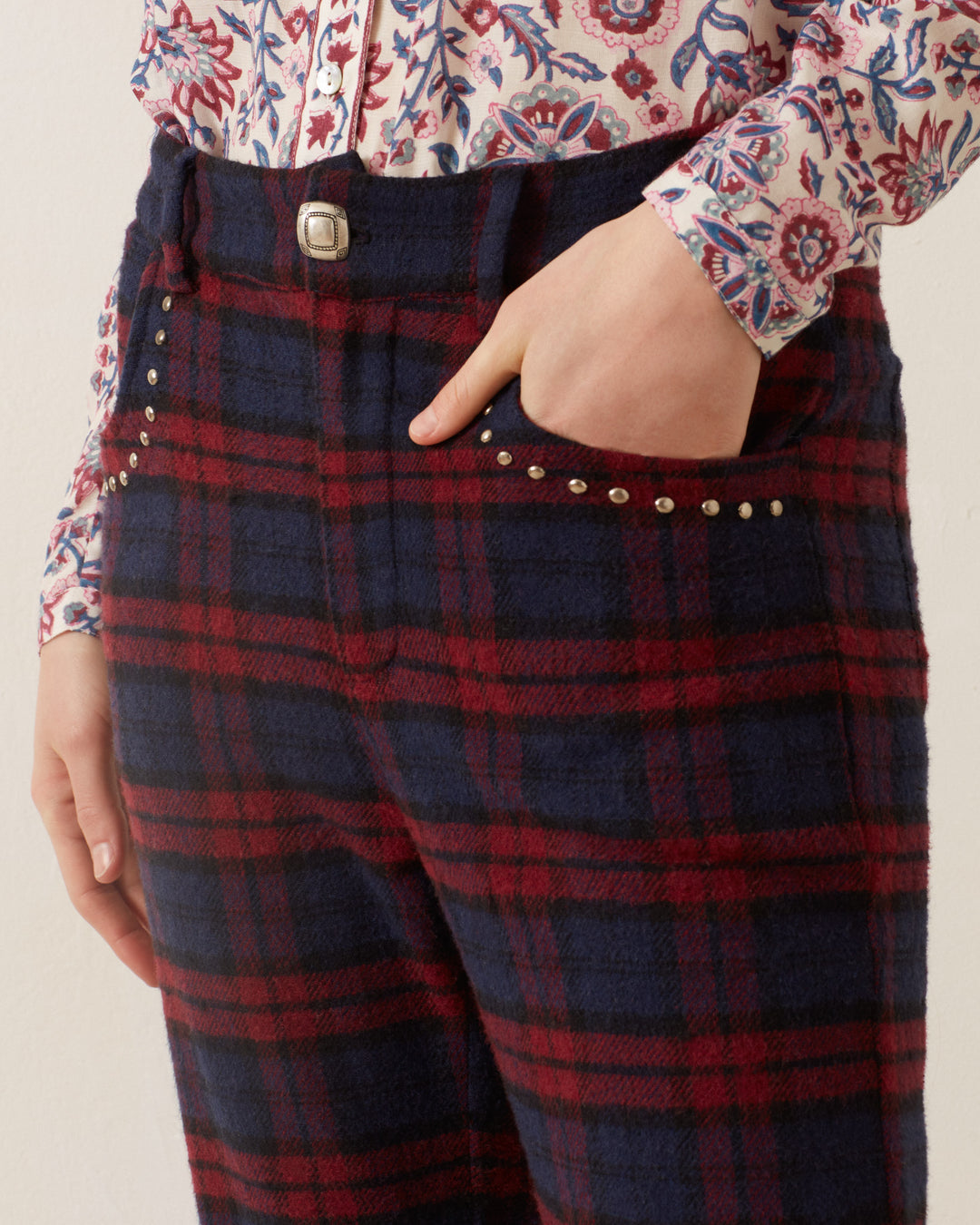 A person is shown wearing the Bunny Berry Plaid Pant, which features silver studs along the pocket seam. With one hand in their pocket, they pair these stylish pants with a chic blouse adorned with red, blue, and white floral patterns to complete their ensemble.