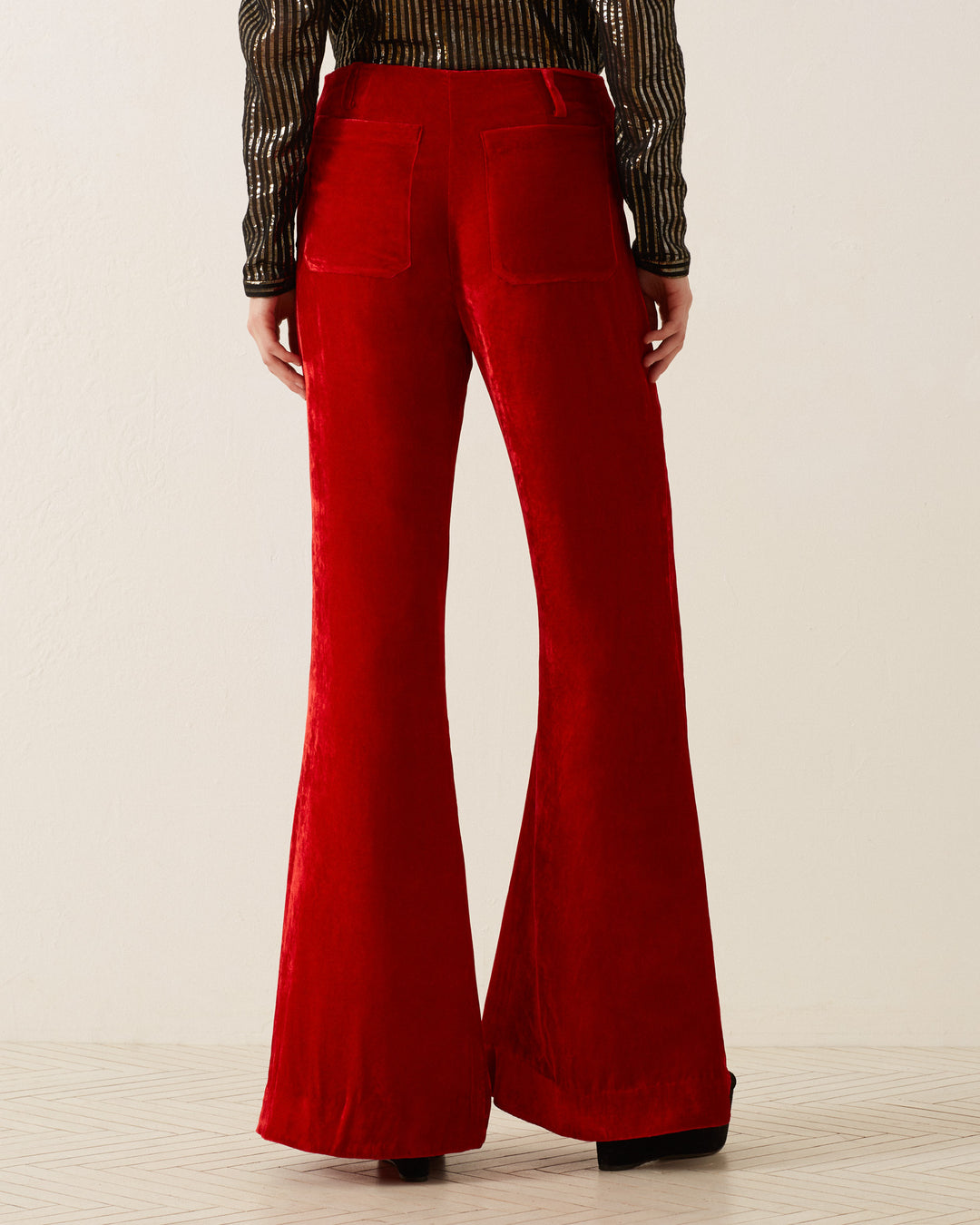 A person is wearing the 'Charlie Cherry Velvet Pant,' featuring a '70s-style flared design in red velvet, paired with a long-sleeve, striped black and gold top. They are standing on a light-tiled floor, with the view from behind highlighting the trousers' high waist and two back pockets.