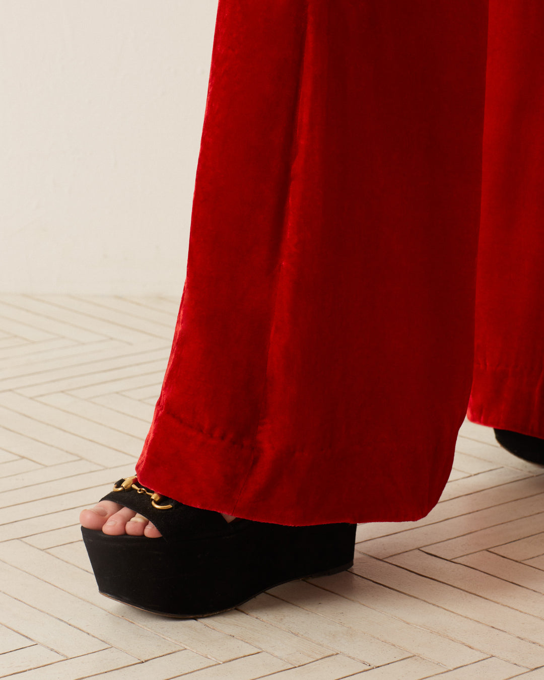 Close-up of a person wearing the 'Charlie Cherry Velvet Pant,' showcasing its '70s-style red velvet and bell-bottom flare, paired with black platform sandals featuring a gold chain detail. The floor beneath showcases a light-colored, herringbone pattern.