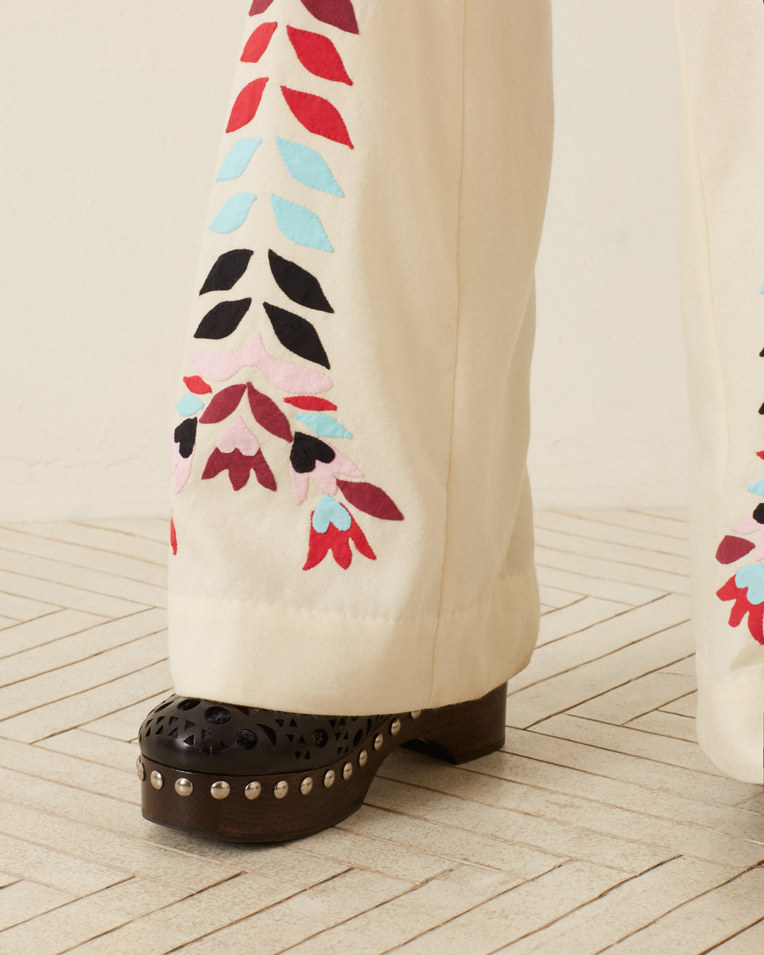 Close-up of the Charlie Ivory Prism Pant leg, showcasing colorful floral appliqué reminiscent of '70s style, paired with a black shoe featuring decorative cutouts and heel embellished with silver studs, all placed on a light wood floor.