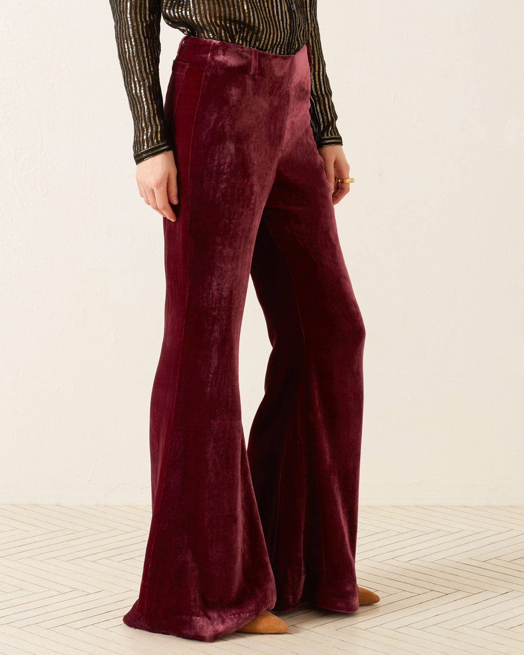 A person embodies '70s fashion in Charlie Plum Velvet Pants, coupled with a long-sleeved black and gold striped top. They are positioned on a light herringbone-patterned floor against an off-white wall, with brown pointed shoes subtly visible.