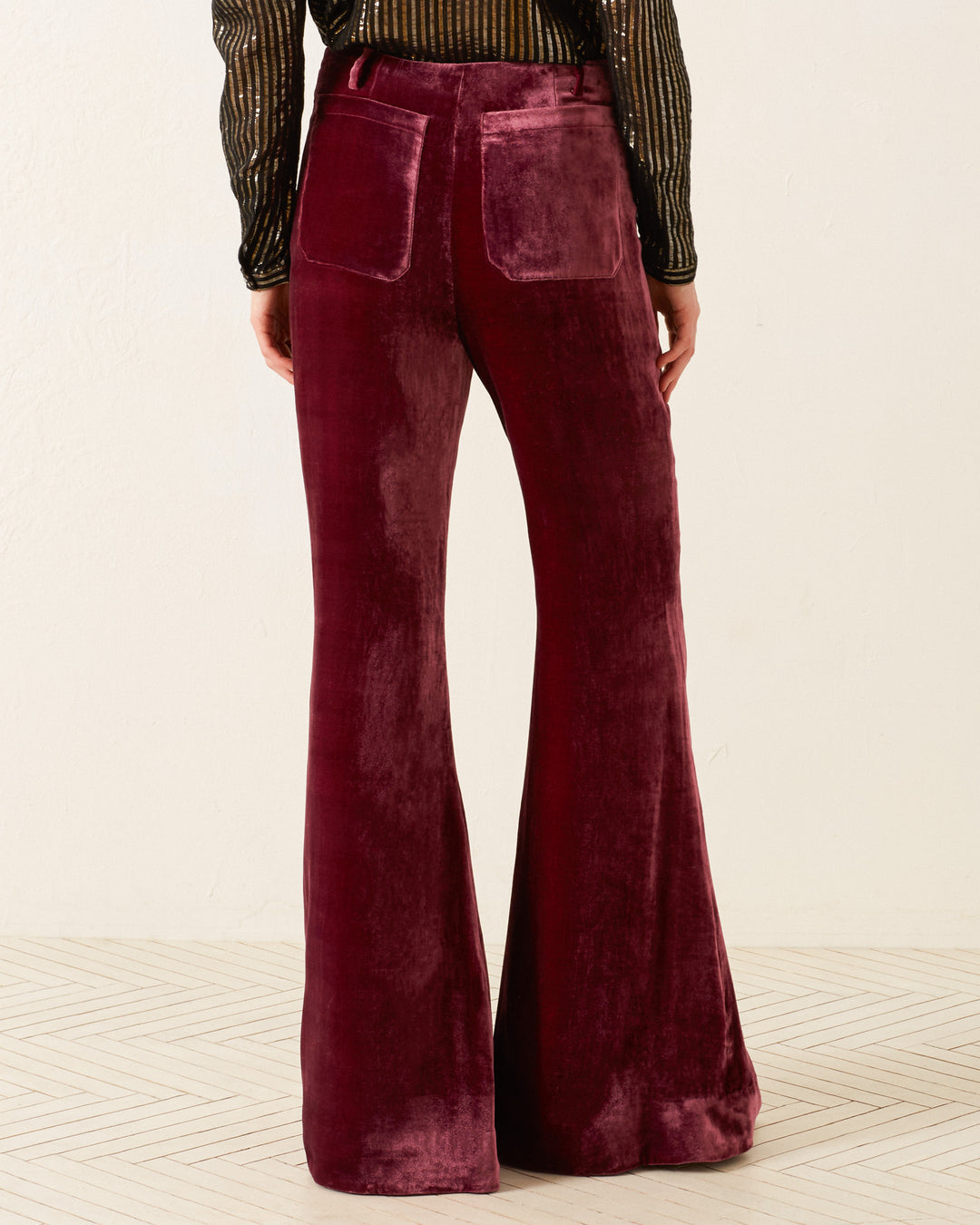 A person wearing the 'Charlie Plum Velvet Pant,' a pair of '70s style velvet bell-bottom pants with large back pockets, stands with their back facing the camera. They are also donning a long-sleeved black and gold striped top, which perfectly complements the retro vibe against a light-colored wall and floor.