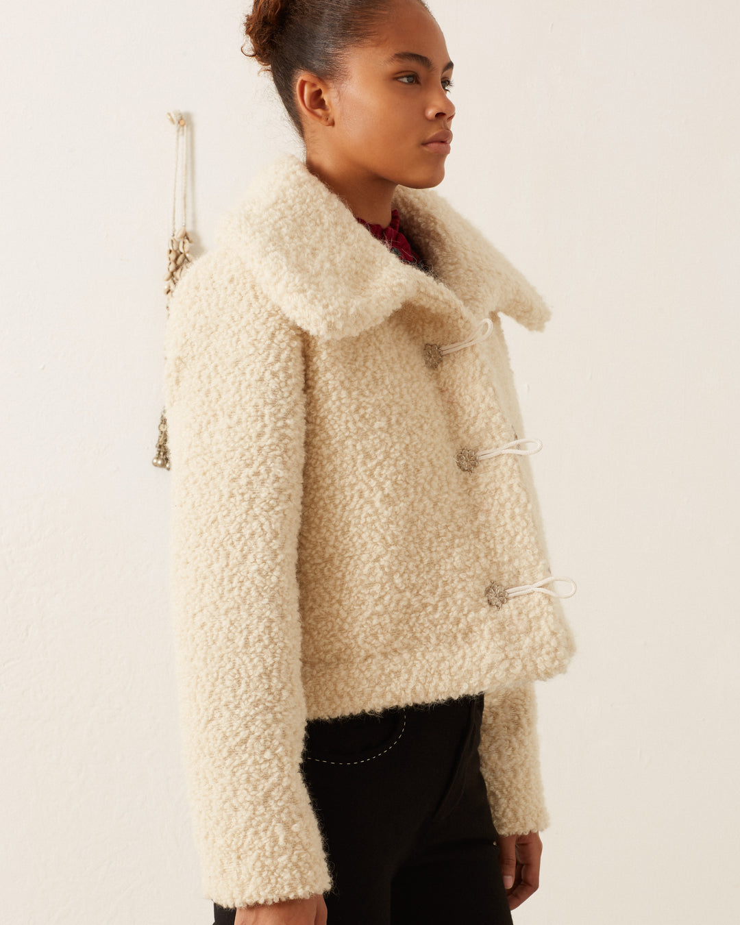 Against a simple backdrop, a woman models the Colby Suri Wool Jacket, featuring a cream hue, textured design, toggle closures, and an oversized collar. Her hair is styled up as she gazes sideways, seamlessly complementing the jacket with dark pants.