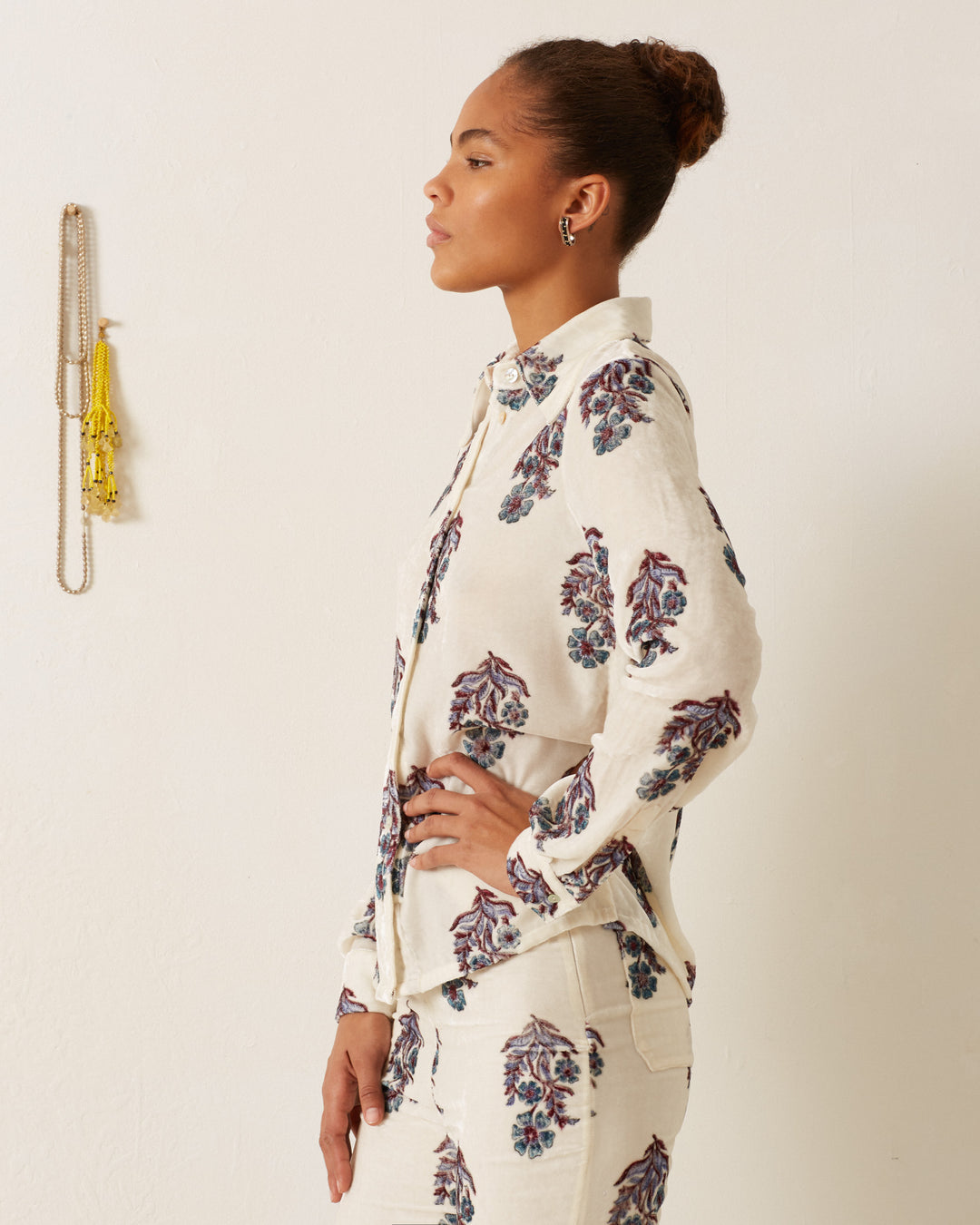 A woman wearing the Dolly Azure Mirabilis Shirt paired with pants stands in profile against a white wall adorned with hanging jewelry.