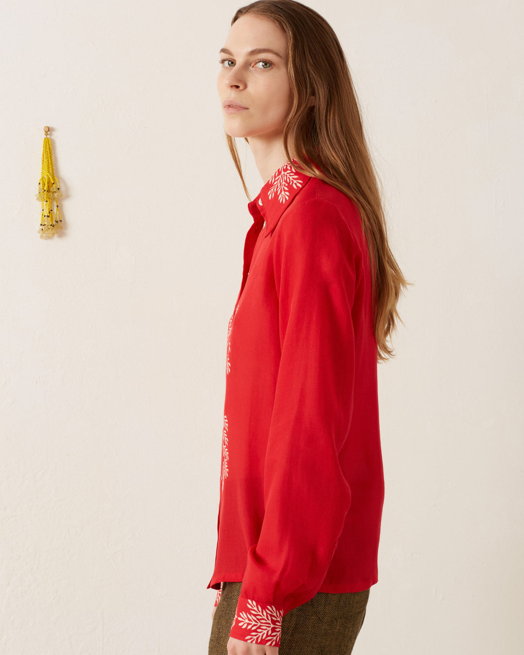 A person with long brown hair is wearing the vintage-inspired Dolly Cherry Mint Shirt, which features white embroidery on the collar and cuffs. They stand sideways, looking forward against a plain off-white wall adorned with a small, decorative gold tassel.