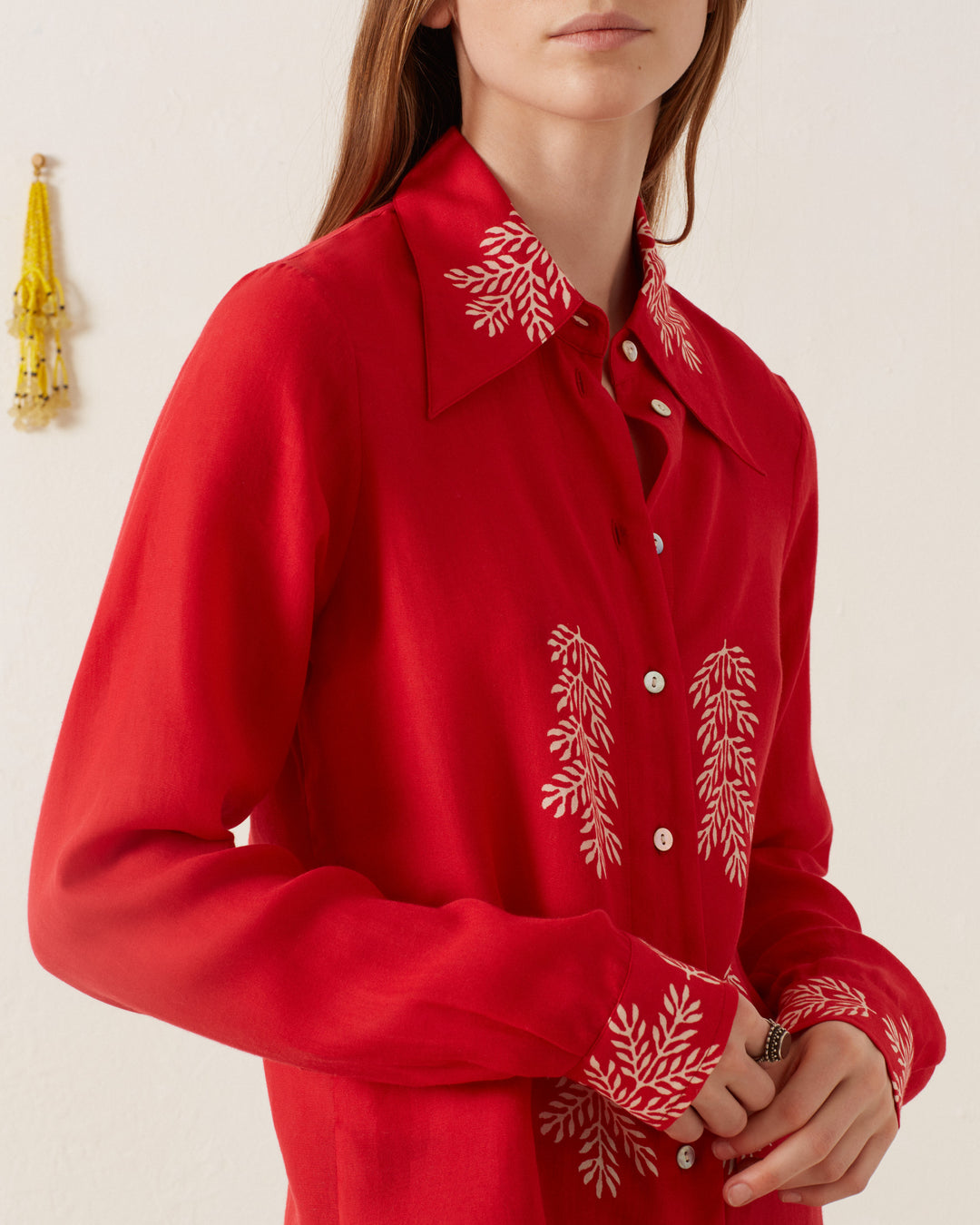 A person wearing a Dolly Cherry Mint Shirt, featuring white embroidered patterns on its vintage-inspired red fabric at the collar, chest, and cuffs, stands against a light wall adorned with a small ornamental tassel. The person's face is not visible.