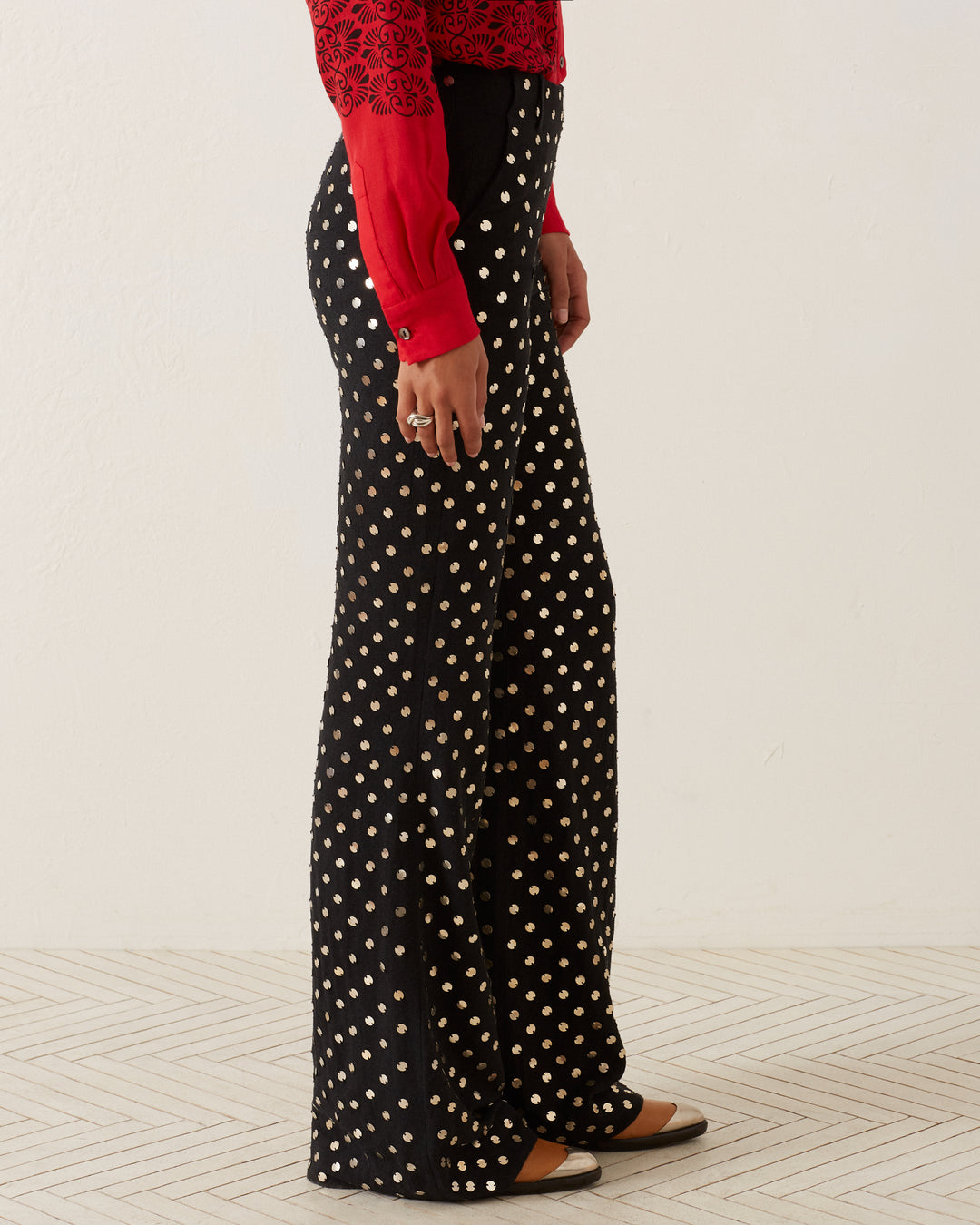 A person wearing Charlie Stardust Pant embellished with silver sequins and a red blouse is shown in profile. They are standing against a light background, their feet adorned in black and gold shoes.