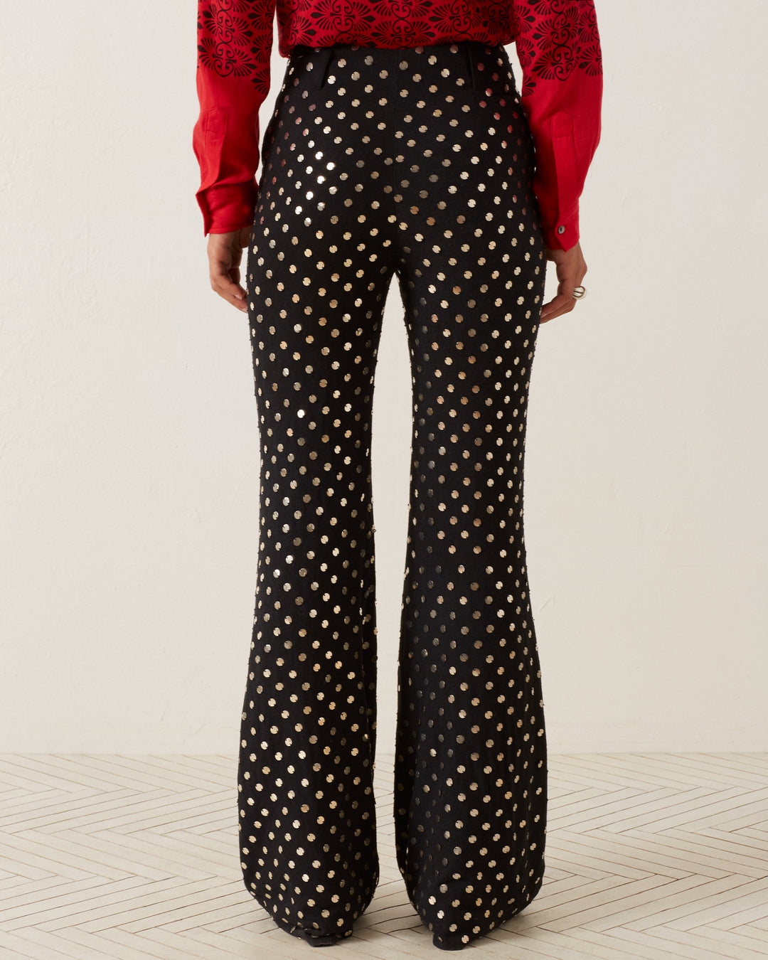 A person is standing on a light-colored, textured silk floor, wearing the Charlie Stardust Pant in black with gold polka dots and a red long-sleeve top. The image focuses on their lower back and legs.