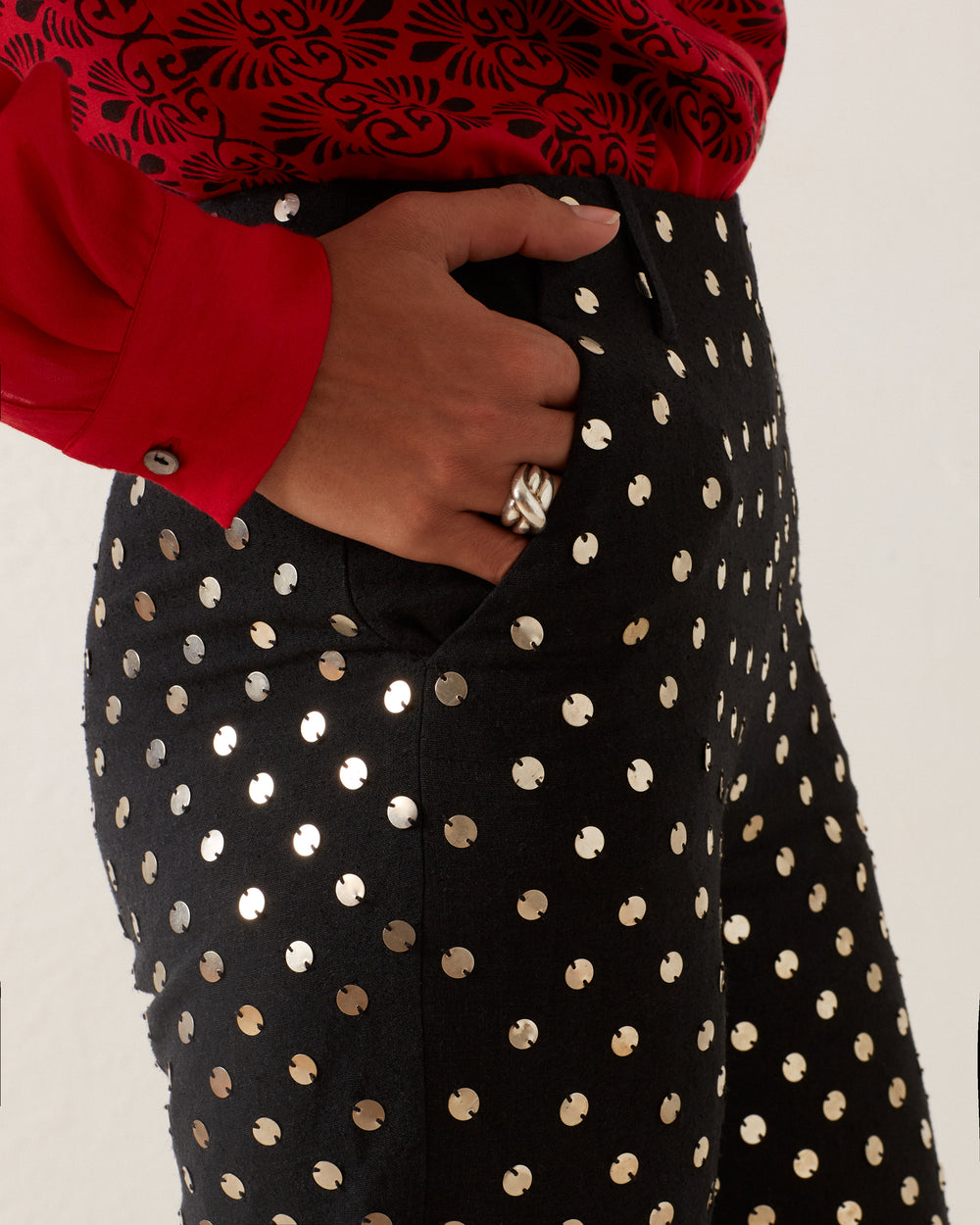 Close-up of a person wearing a red long-sleeve shirt with the Charlie Stardust Pant, adorned with shiny circular studs and silver sequins. The person’s hand, wearing a silver ring, is partially tucked into the pants pocket.