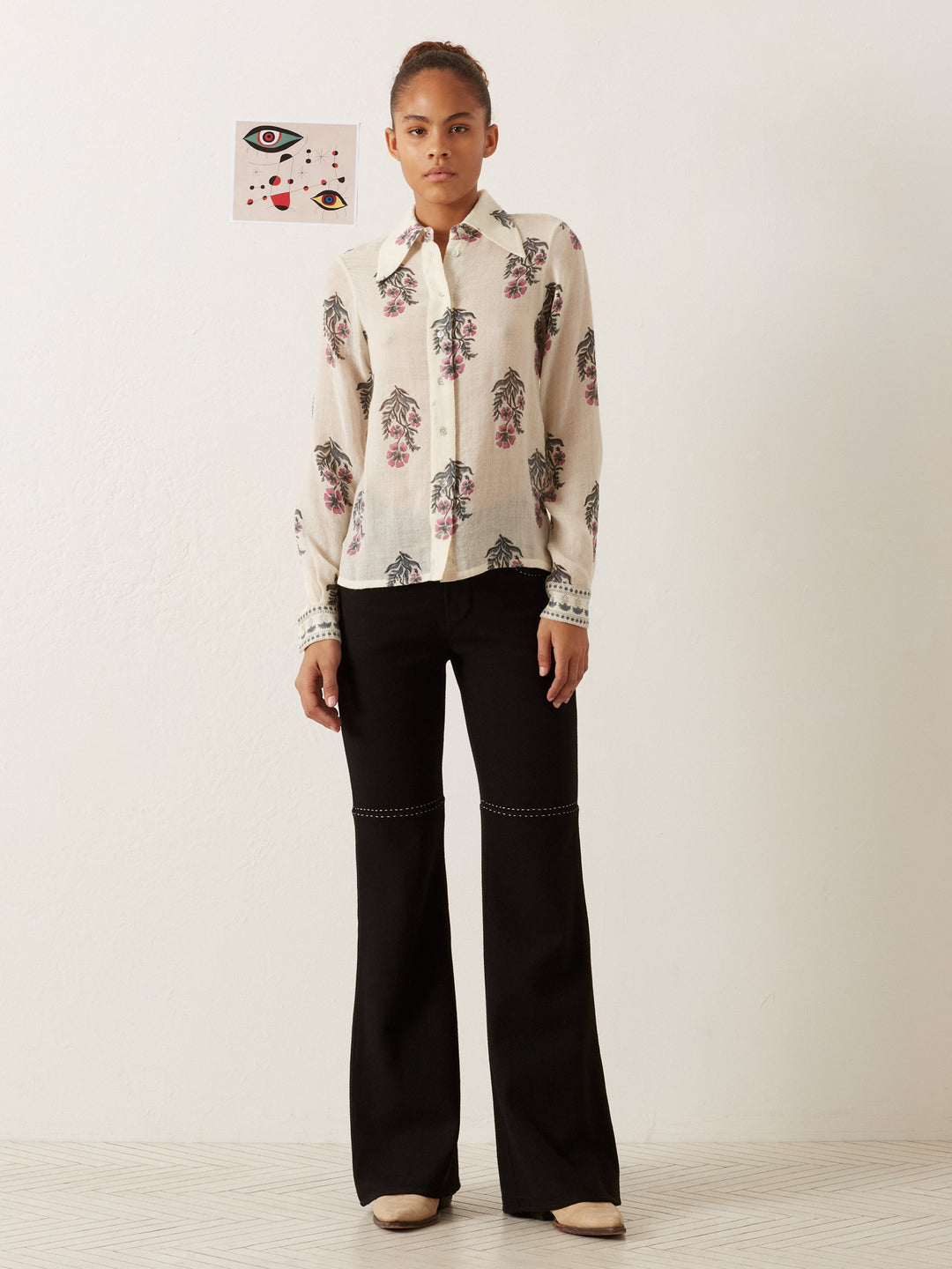 A person stands against a plain wall, wearing the Dolly Mirabilis Wool Shirt with its vintage-inspired floral print and black wide-leg pants. Beige shoes complete the outfit. A small, abstract artwork featuring eyes is hung on the wall behind them.