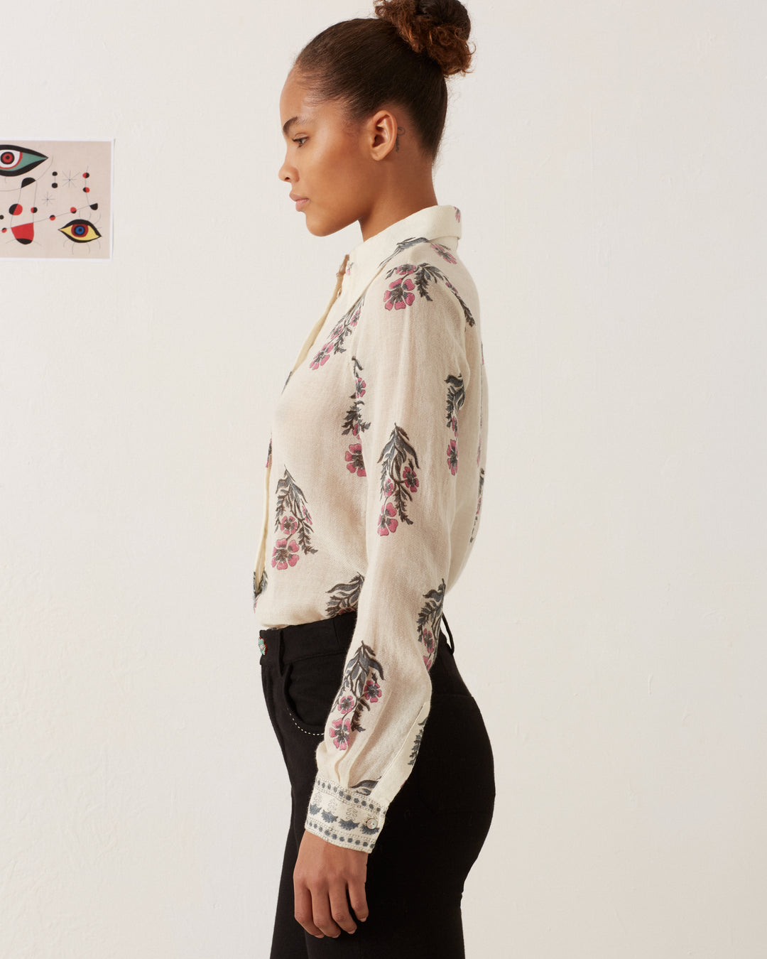 A woman with a bun hairstyle stands in profile against a plain white background, wearing the Dolly Mirabilis Wool Shirt, which is adorned with vintage-inspired floral prints, paired with black pants. A small, abstract face artwork is attached to the wall behind her.