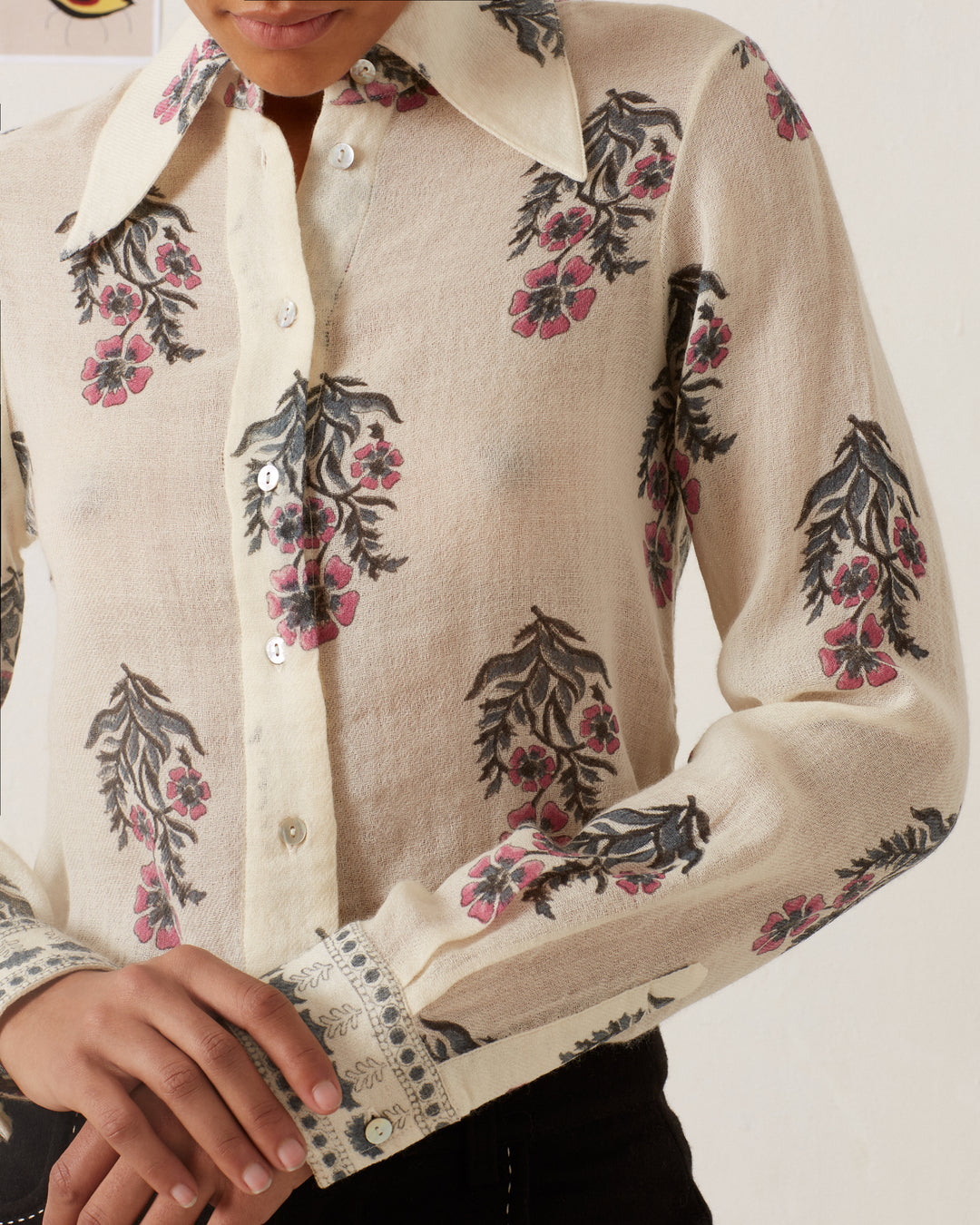 A person is wearing the Dolly Mirabilis Wool Shirt in beige, featuring long sleeves with a vintage-inspired floral motif of pink and black flowers. The shirt has a pointed collar, button-down front, and decorative cuffs. The individual is shown from the neck down, with their left hand resting near the waist.