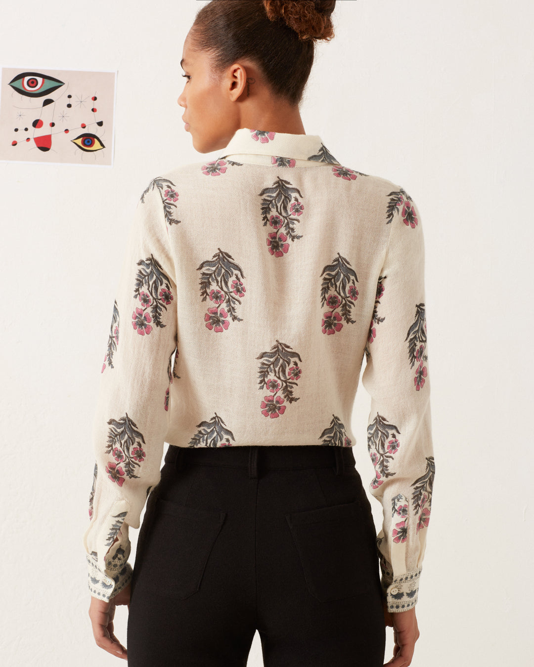 A person with a bun hairstyle is facing away, wearing a Dolly Mirabilis Wool Shirt with a floral print and black trousers. On the wall in front of the person, there is an abstract art piece featuring eyes and geometric shapes.