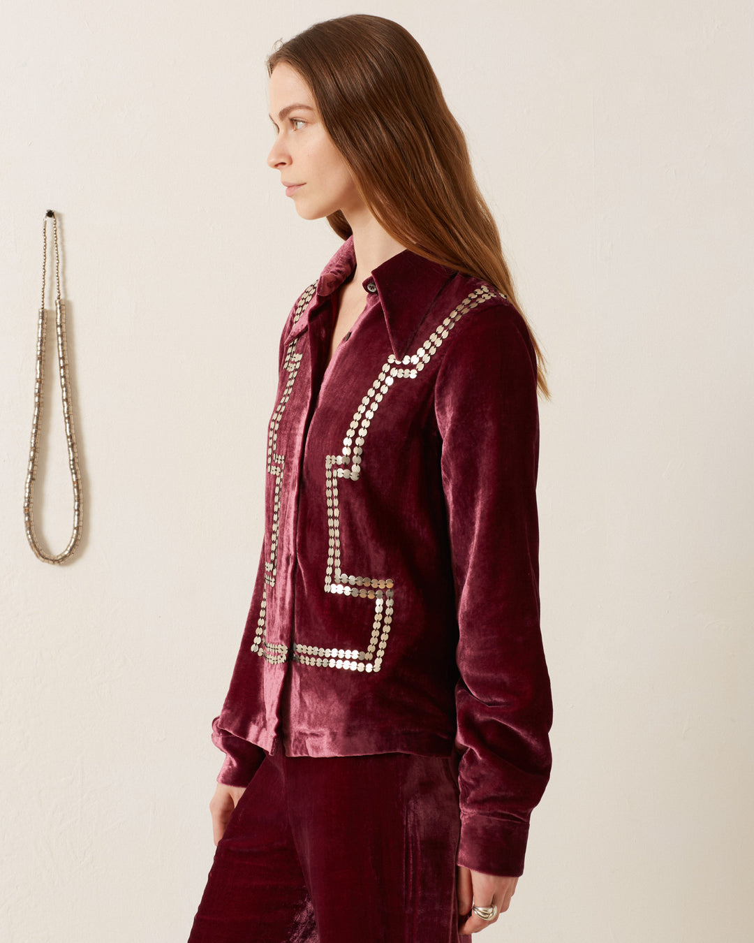 A person with long hair wearing the Dolly Plum Velvet Shirt, which is vintage-inspired and adorned with decorative patterns, stands in profile view. A silver chain hangs on the wall.