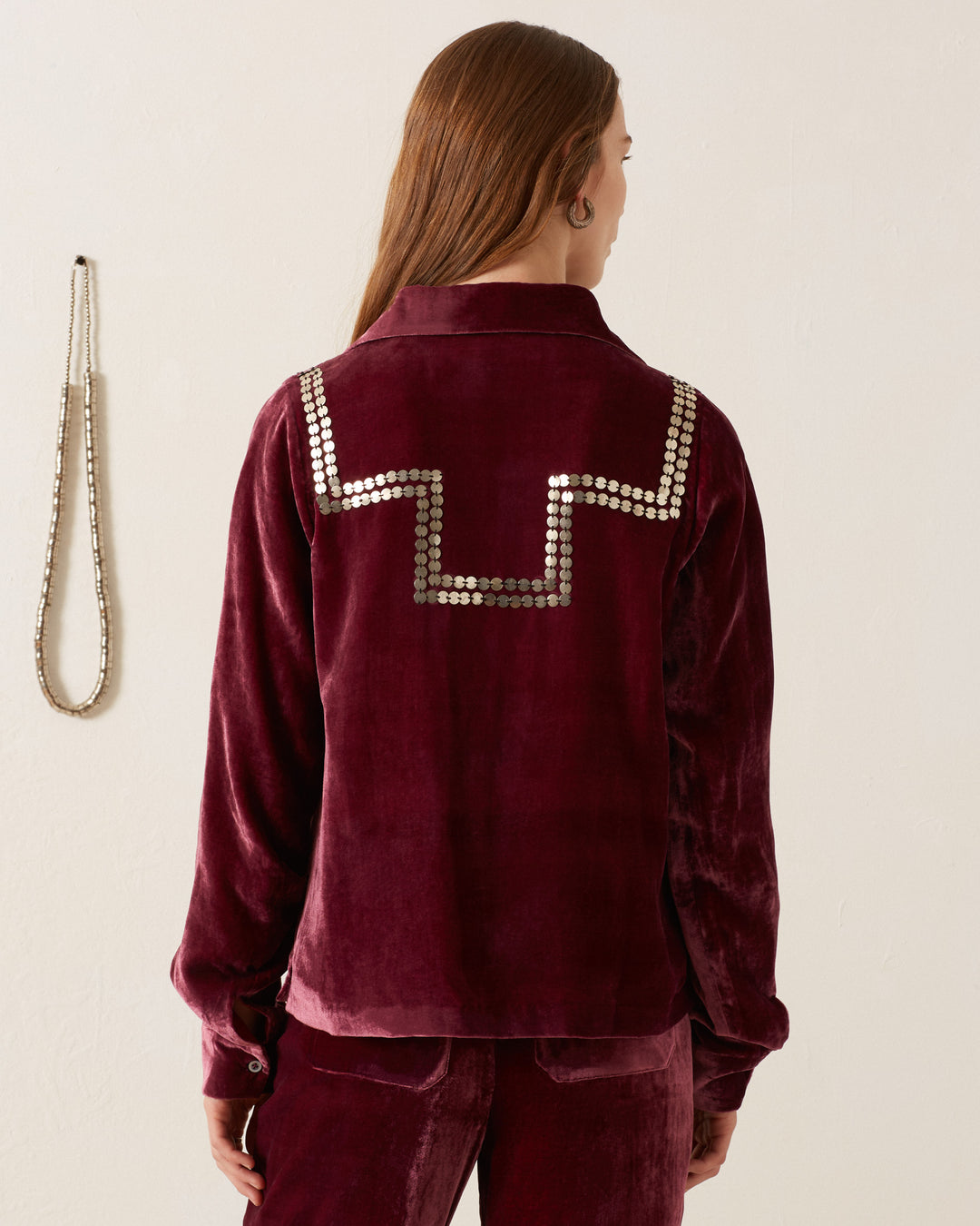 The person is facing away, wearing the Dolly Plum Velvet Shirt in burgundy, adorned with metallic embellishments on the back. Their hair is loose against a neutral background, with a necklace hanging on the wall.